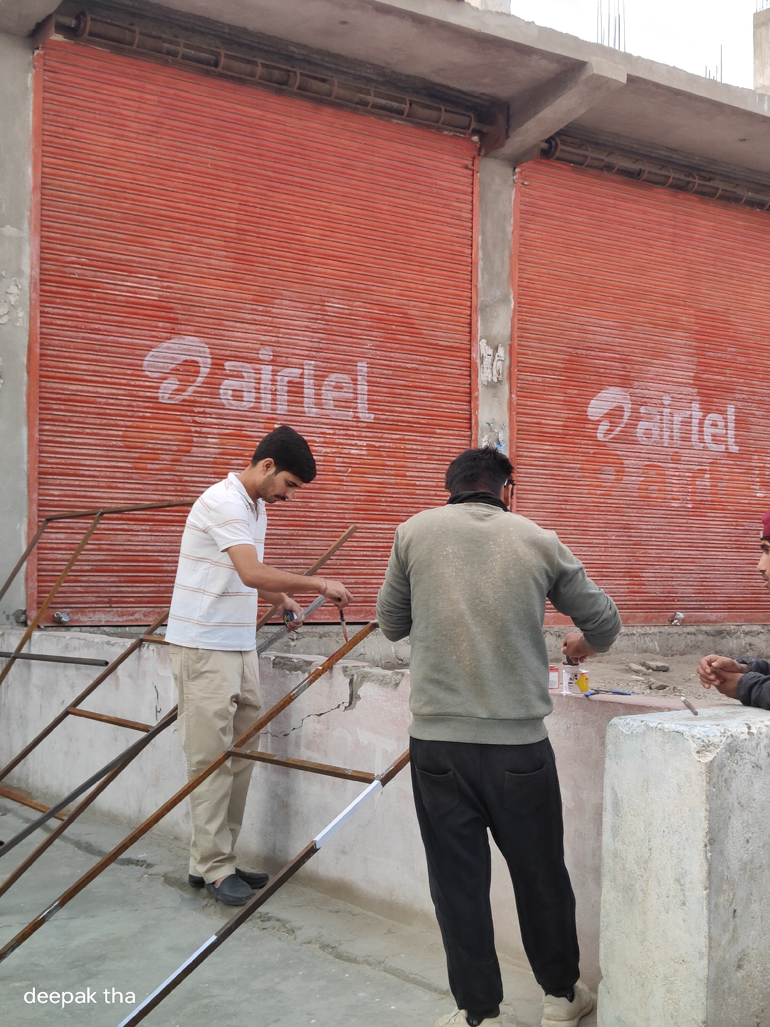 A construction site in Leh, India, features a building with two large, red, corrugated metal roll-up doors, each displaying a white "airtel" logo. In the foreground, two men are engaged in construction work. The man on the left, wearing a striped t-shirt and beige pants, is holding a tape measure and a brush, appearing to be marking or painting a metal frame. The man on the right, in a grey sweatshirt and black pants, is holding a small container and a brush, possibly working on a similar task. A third person, partially visible on the far right, has their hands clasped. The ground is a rough concrete surface, and scaffolding or metal framing structures are present. The overall environment suggests an outdoor, possibly urban, construction setting.