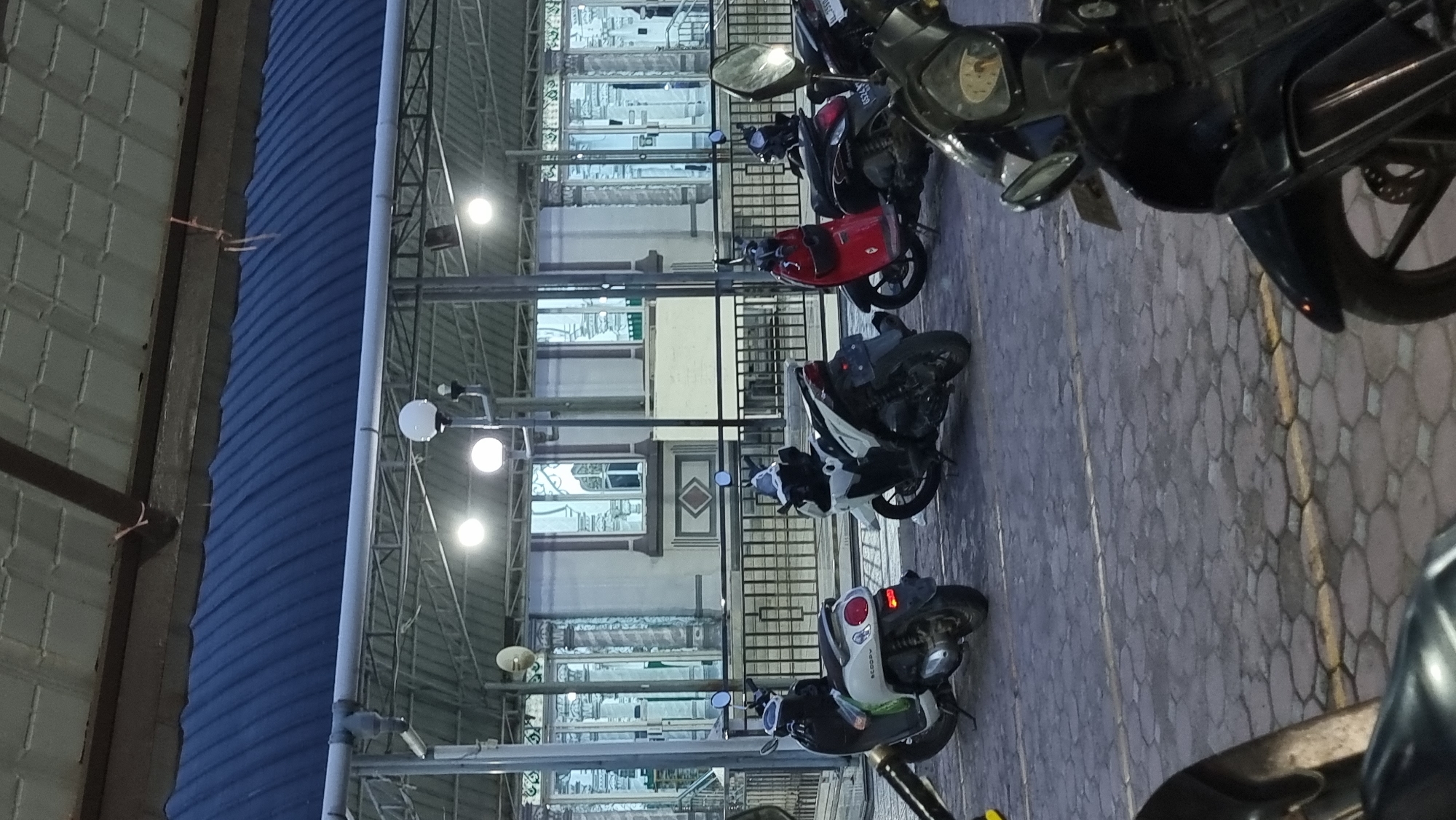 In Keudekruenggeukueh, Indonesia, a nighttime scene unfolds in front of a building with reflective surfaces and ornate architectural details, possibly a mosque or a public institution. A covered outdoor area with a metal roof structure and exposed beams shelters a row of parked motorcycles. The ground is paved with interlocking hexagonal stones, marked by white and yellow lines. Several scooters are visible, including a white one on the left with the "Scoopy" logo, a white and black one in the center, and a red one to its right. The lighting appears to be from overhead fixtures, casting a glow on the scene. The building in the background has large, mirrored windows that reflect the surrounding lights and structures. The overall impression is one of a public space utilized for parking, likely at the end of the day or during evening hours.