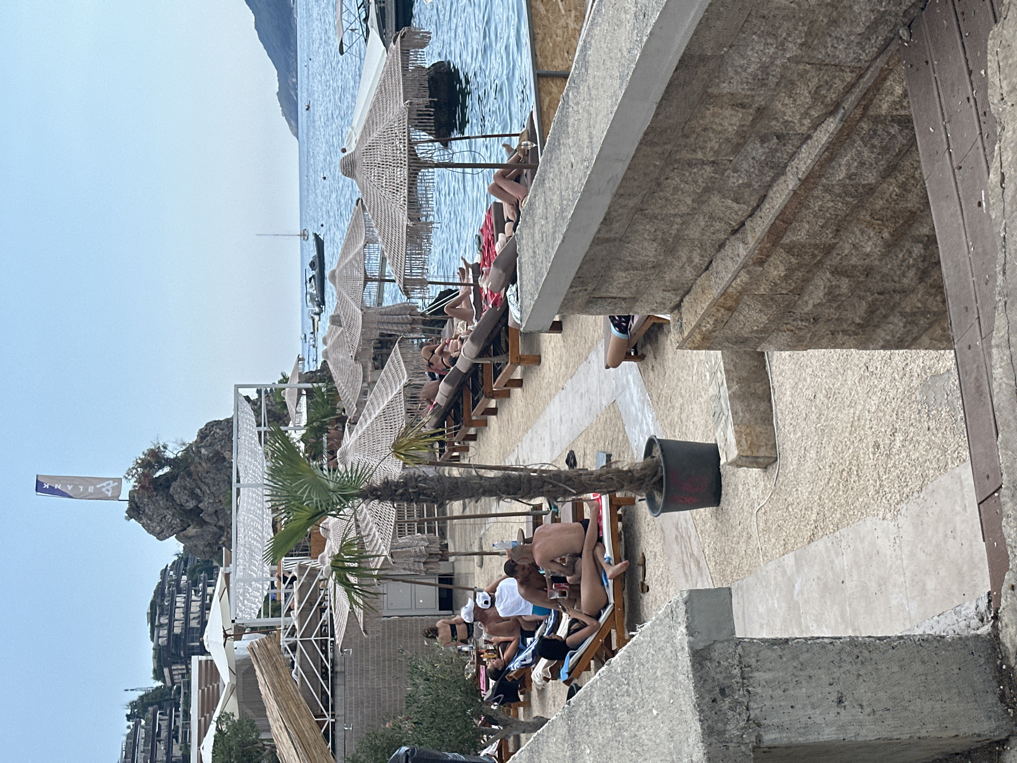 The image depicts a beachside setting in Budva, Montenegro, characterized by a bustling beach club and a clear blue sky. In the foreground, a concrete wall and steps lead down to a sandy area populated with sunbathers lounging on chairs under straw umbrellas. Palm trees are scattered throughout the scene, with one prominently featured in a large black pot.
Several groups of people are visible, some engaging in conversation while others relax or sunbathe. One group of men is seated around a table, appearing to be enjoying drinks. The background reveals the turquoise waters of the Adriatic Sea, dotted with a few boats, and a verdant, mountainous coastline. Buildings are perched on the hills overlooking the beach, and a distinctive rock formation with a flag stands near the water's edge. The overall atmosphere suggests a vibrant and popular tourist destination.