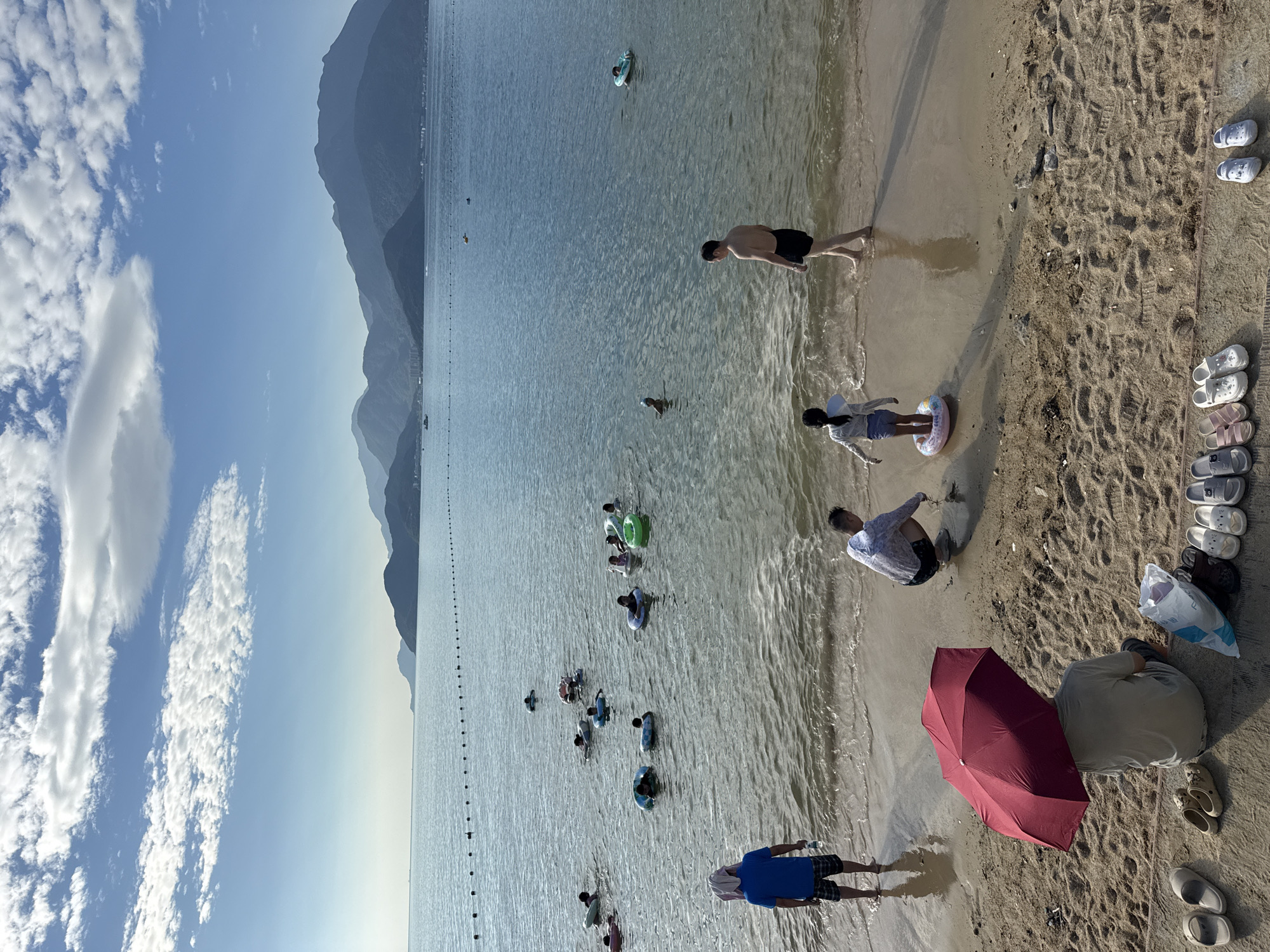 In the city of Longgang District, China, a scenic beach scene unfolds under a blue sky dotted with white clouds. The calm, clear ocean stretches to the horizon, where it meets a range of distant mountains. A floating rope barrier demarcates a swimming area in the water, within which numerous people are seen enjoying themselves. Some individuals are floating on colorful inflatable rings, while others are swimming or wading in the shallow water.
Closer to the shore, a group of people are gathered on the sandy beach. In the foreground, a person is seated under a large red umbrella, providing shade. Adjacent to them, a collection of various flip-flops and sandals are neatly arranged. Nearby, another person is crouched down, appearing to examine something on the sand. Further down the beach, a man in black shorts is walking towards the water, and a young girl wearing a swimsuit and a life vest is standing in the shallow water, seemingly preparing to enter the sea. On the left side of the frame, a man in a blue t-shirt and plaid shorts stands facing the ocean, holding a bottle of water. The overall atmosphere is one of leisure and outdoor recreation.