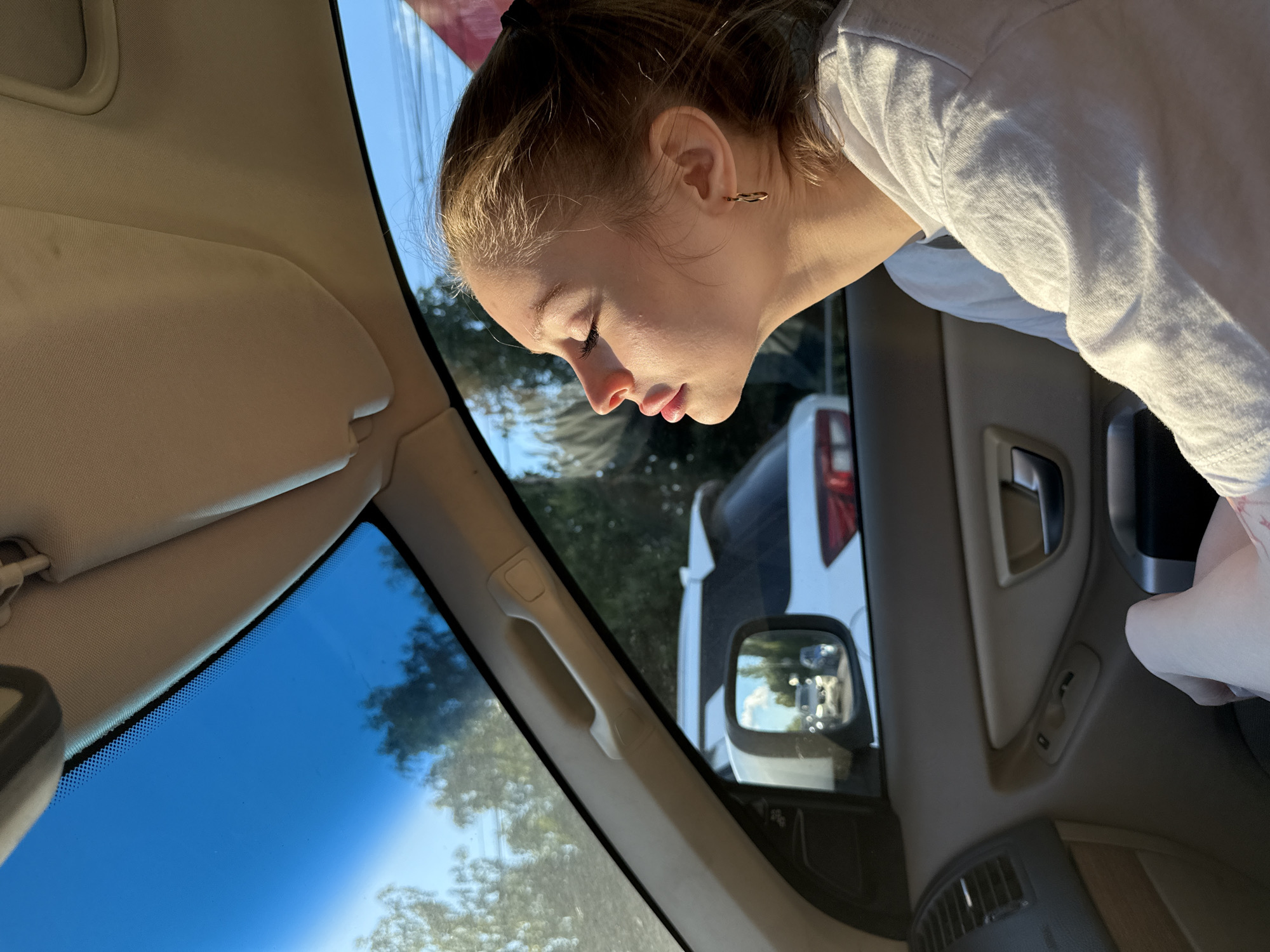 A young female, likely a teenager or young adult with light brown hair tied back, is captured leaning out of a car window, looking intently downwards. Her face is in profile, illuminated by strong, warm sunlight, indicating it is daytime with a clear sky. She is wearing a light-colored (possibly white or light grey) short-sleeved t-shirt and has a small hoop earring in her visible ear.
The scene is set inside a car, looking out the passenger side. The car's interior features a beige/tan headliner, a sun visor, and an A-pillar with a grab handle. Part of the passenger door panel, showing controls (likely for windows or mirrors), and a dashboard vent are also visible.
Outside the car, green trees and foliage are seen against a bright blue sky. A white vehicle, possibly an SUV or van, is visible in the background, and its reflection can be seen in the car's side mirror. The overall impression is one of focused observation or curiosity. No discernible text is visible in the image. The provided context indicates the location is Vniissok, Russia.