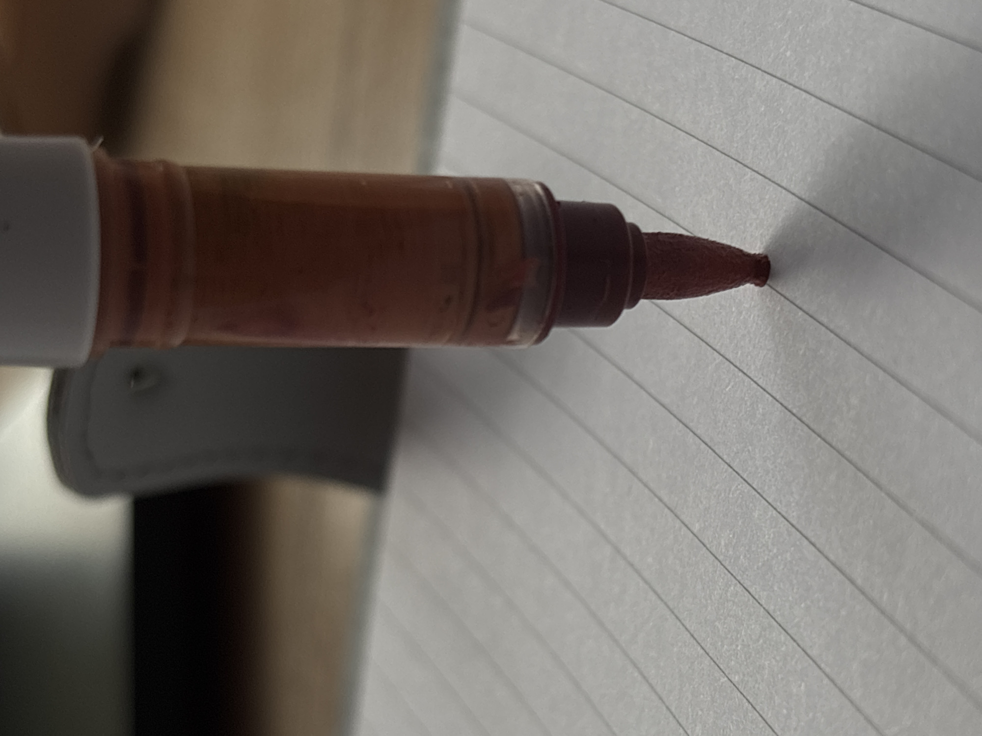 A close-up, low-angle shot shows the tip of a red marker pen making contact with lined notebook paper. The marker has a translucent barrel with visible red ink inside. The paper is white with evenly spaced gray lines. The background is blurred, showing a hint of a gray object and a wooden surface. There are no people or other objects clearly visible. The lighting appears to be natural, creating a soft shadow from the marker tip. The scene suggests a moment of writing or drawing.