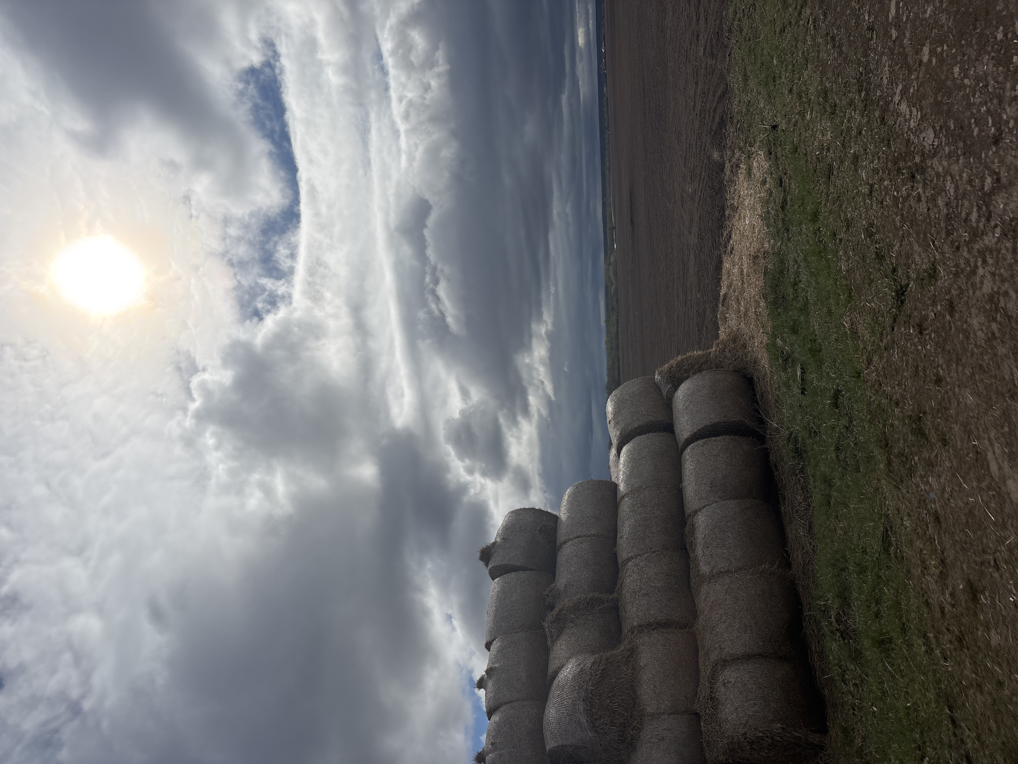 The image shows a rural landscape in Mokhnutino, Russia. A stack of round hay bales is in the foreground, positioned on a field alongside a patch of dry grass. Beyond the hay bales, the landscape transitions into a plowed field. Above, a dramatic sky filled with clouds is visible, with the sun shining brightly through a gap in the cloud cover.