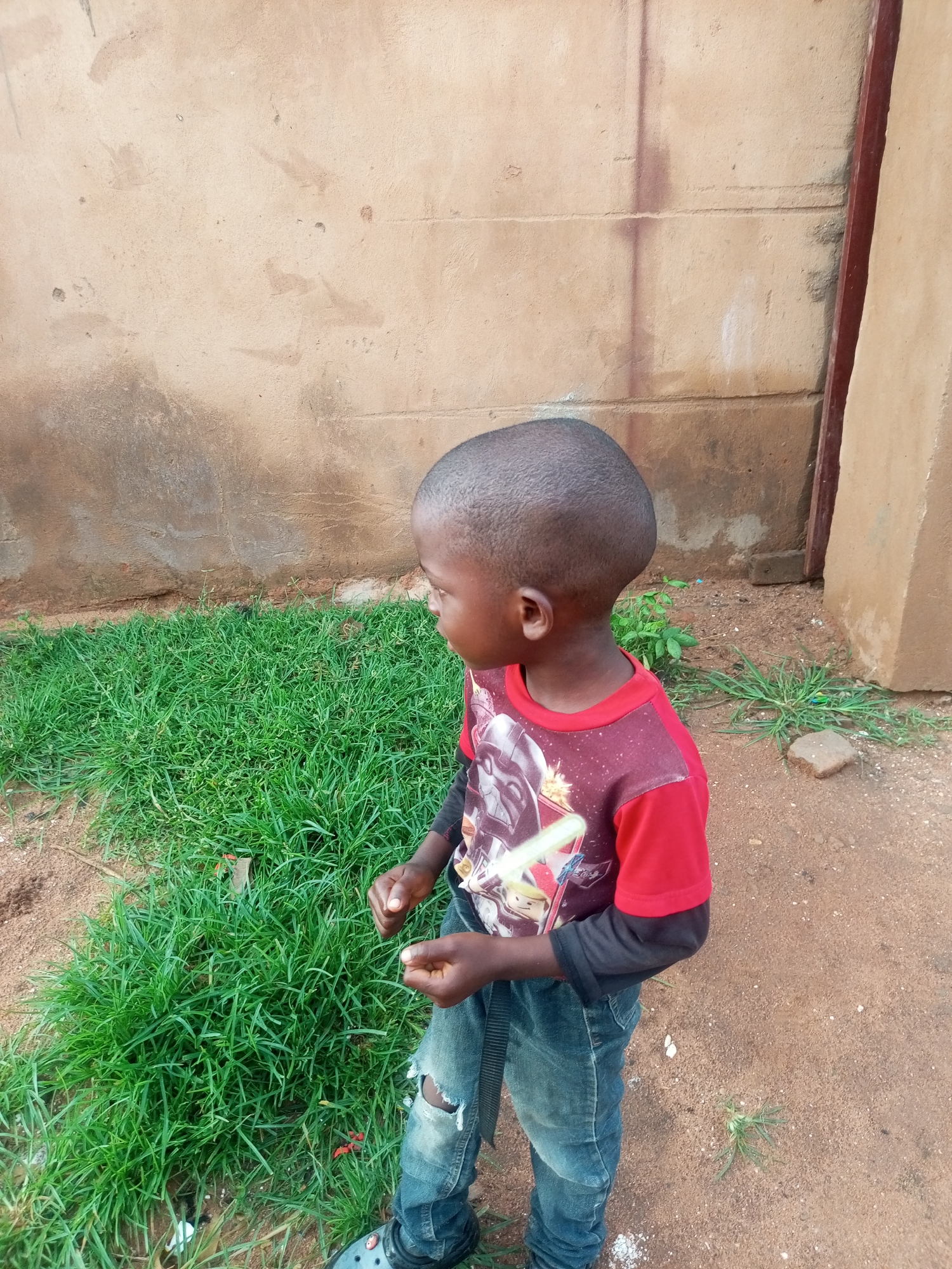 A young boy stands outdoors near a dirt path and a patch of lush green grass, with a beige, textured wall forming the background. He is wearing a maroon t-shirt with a graphic print and black long sleeves, paired with distressed blue jeans. His hands are held in front of him, and he is looking to his left, with his head turned slightly. The scene appears to be in Tempure, Nigeria.