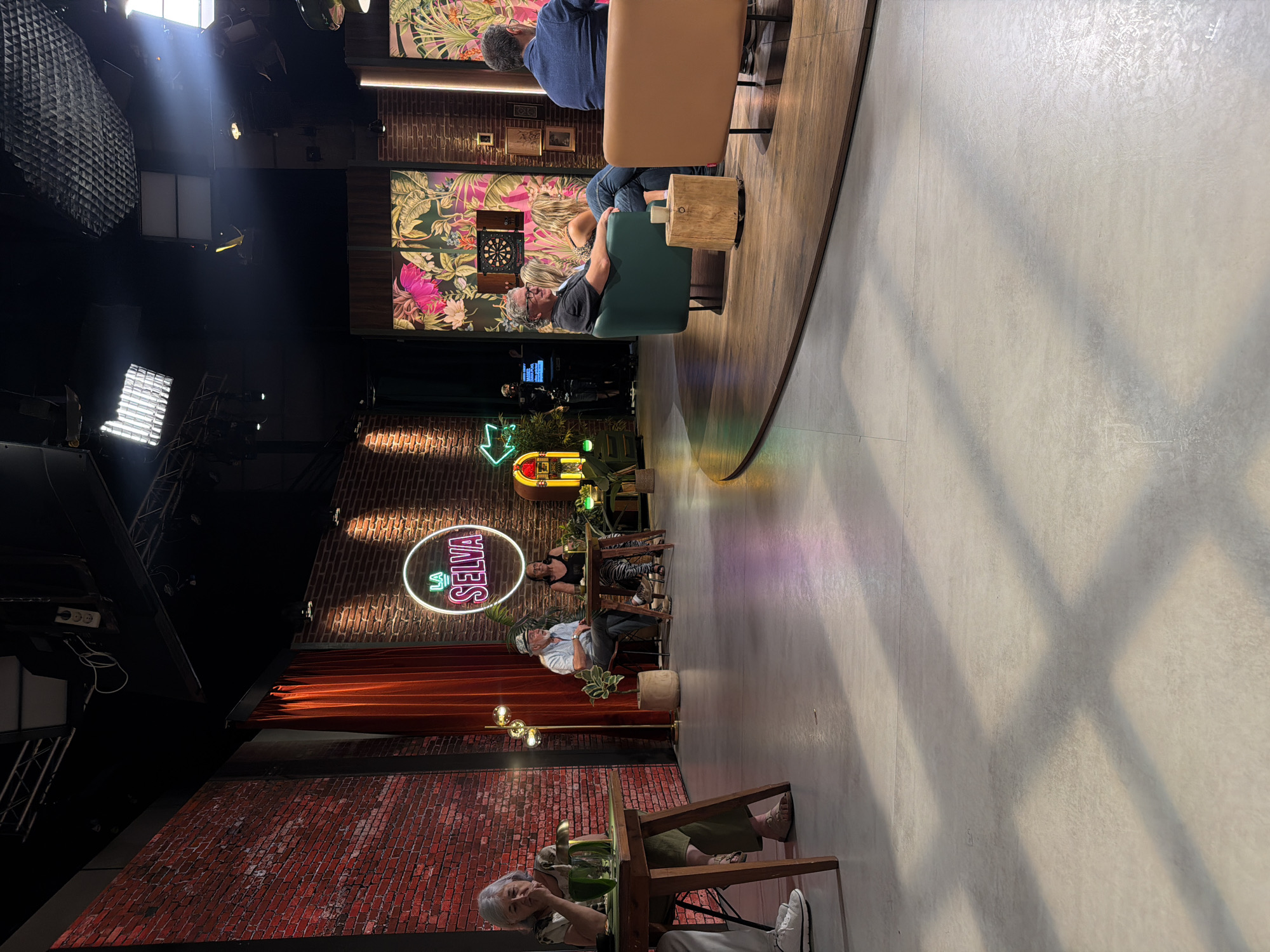 The image depicts a television studio set in Sant Joan Despí, Spain, likely for a show called "La Selva." The set features a brick wall backdrop with a circular neon sign that reads "LA SELVA." In the foreground, several people are seated at tables and on chairs, appearing to be guests or participants in a broadcast. One woman in a light-colored top and green pants sits at a table on the left, with a glass of water in front of her. Further back, a man in a hat and a woman in a dark top are seated at another table. To the right, a man in a grey t-shirt and a woman in blue jeans are seated on a green sofa. Another person in a blue jacket is visible in the background. The studio is equipped with lighting rigs and decorative elements such as plants and a vintage-style jukebox. The overall atmosphere suggests a casual and engaging television program.
