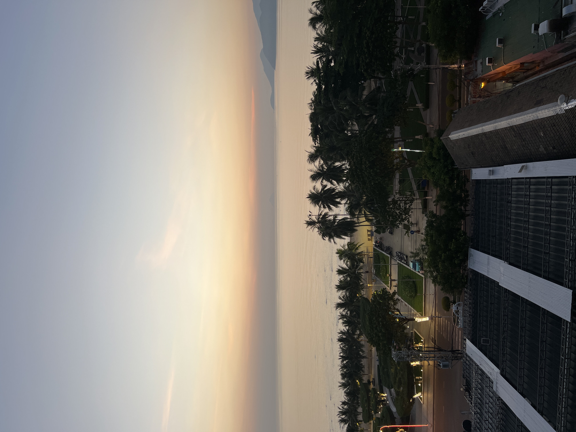 This is an aerial photograph of a beachside scene in Nha Trang, Vietnam, at dawn or dusk. The sky transitions from a pale blue at the top to soft orange and yellow hues closer to the horizon. The calm sea stretches out, reflecting the muted colors of the sky. A line of palm trees borders the beach, with a park-like area featuring manicured lawns and walkways visible behind them. Streetlights and some decorative lighting are illuminated, suggesting the early morning or late evening. Several motorcycles are parked in the area, and a few indistinct figures can be seen walking on the paths. In the foreground, the dark, corrugated metal roofs of buildings are visible, indicating an urban or commercial area adjacent to the beach. The overall impression is one of tranquility and the gentle transition of light.