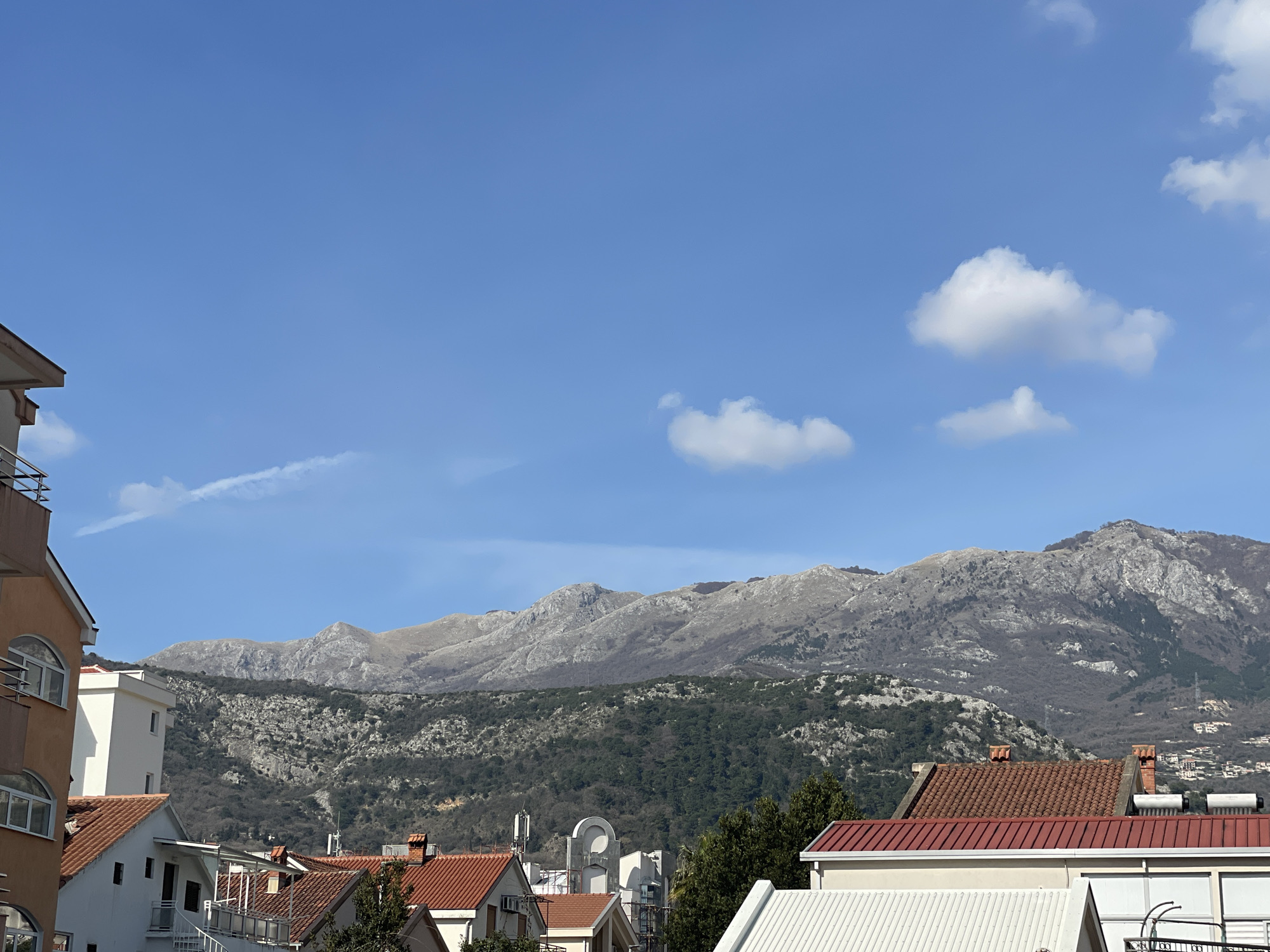 The image displays an urban and mountainous landscape in Budva, Montenegro, observed under a clear blue sky.
In the foreground, a collection of residential buildings is visible from an elevated vantage point. On the far left, a multi-story structure with a light-toned facade features multiple arched windows and balconies equipped with dark metal railings. The central and right portions of the foreground contain various buildings, predominantly characterized by reddish-brown terracotta tile roofs and light-colored, mostly white, facades. One white building centrally positioned exhibits a distinctive rounded upper architectural feature. Further to the right, a building with a bright red corrugated metal roof partially obscures another structure featuring a white corrugated metal roof. Detailed elements on the visible rooftops include multiple terracotta-capped chimneys, cylindrical solar water heaters, and various antennas. Sparse trees, some evergreen and some bare, are interspersed among these structures.
The midground is occupied by a substantial hillside covered with a mix of dense evergreen and deciduous vegetation, appearing as a dark green band with intermittent rocky outcrops.
The background is dominated by a range of rugged, grey limestone mountains. These mountains present extensive exposed rock surfaces and possess sparse vegetation, primarily on their lower slopes, with barren peaks. The sky above is uniformly blue, containing several scattered white cumulus clouds, predominantly in the upper right quadrant. A faint, linear contrail is discernable in the upper left portion of the sky. No human figures are present in the scene.