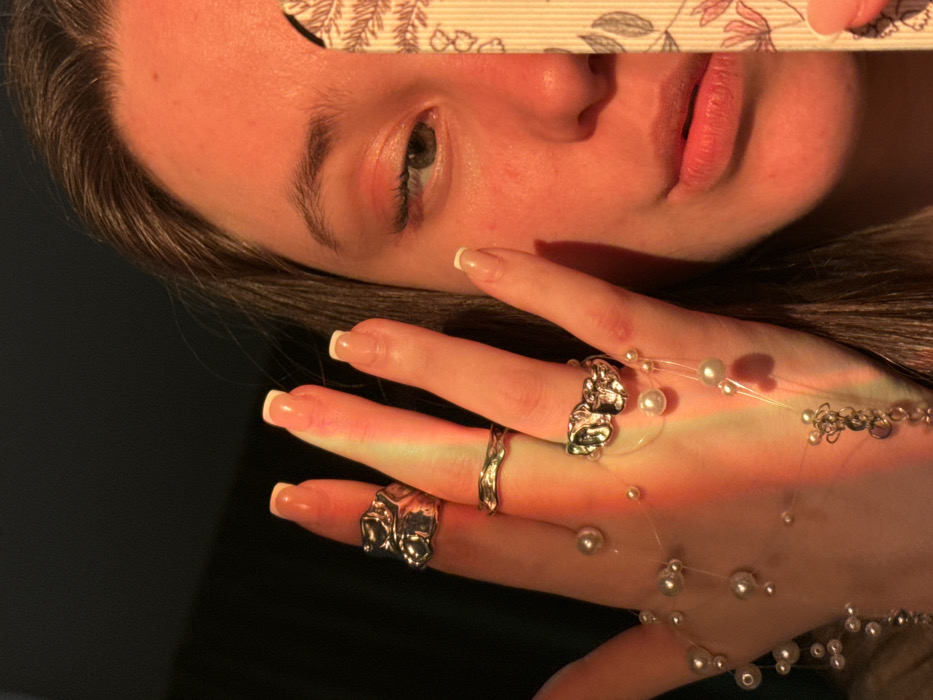 The image presents a close-up, warm-toned portrait of a female individual, primarily focusing on her left hand and the right side of her face. A strong, reddish-orange light source illuminates the scene from the left, casting a glow on her skin and creating a subtle prismatic effect across her fingers. The background is dark and indistinct.
The individual, appearing to be a young adult, has fair skin, dark brown hair partially visible on the right, and a blue-green right eye looking slightly to the left. Her lips are slightly parted. Her fingernails are manicured with white tips.
Her left hand is held forward, displaying three silver-toned rings: an abstract, chunky ring on the index finger, a thin, wavy band on the middle finger, and another abstract, chunky ring on the ring finger. A delicate, multi-strand piece of jewelry, composed of small white pearls and tiny silver beads strung on clear filament, is draped around her fingers and across her palm, with a small cluster of silver charms visible near the base of her hand.
She holds a vertical, rectangular object with her right hand, partially obscuring the lower right portion of her face. This object is light-colored with a repeating pattern of dark botanical elements, resembling a decorative nail file or bookmark.
The specific location within Krasnodar, Russia, cannot be determined from the visual information provided in the image.