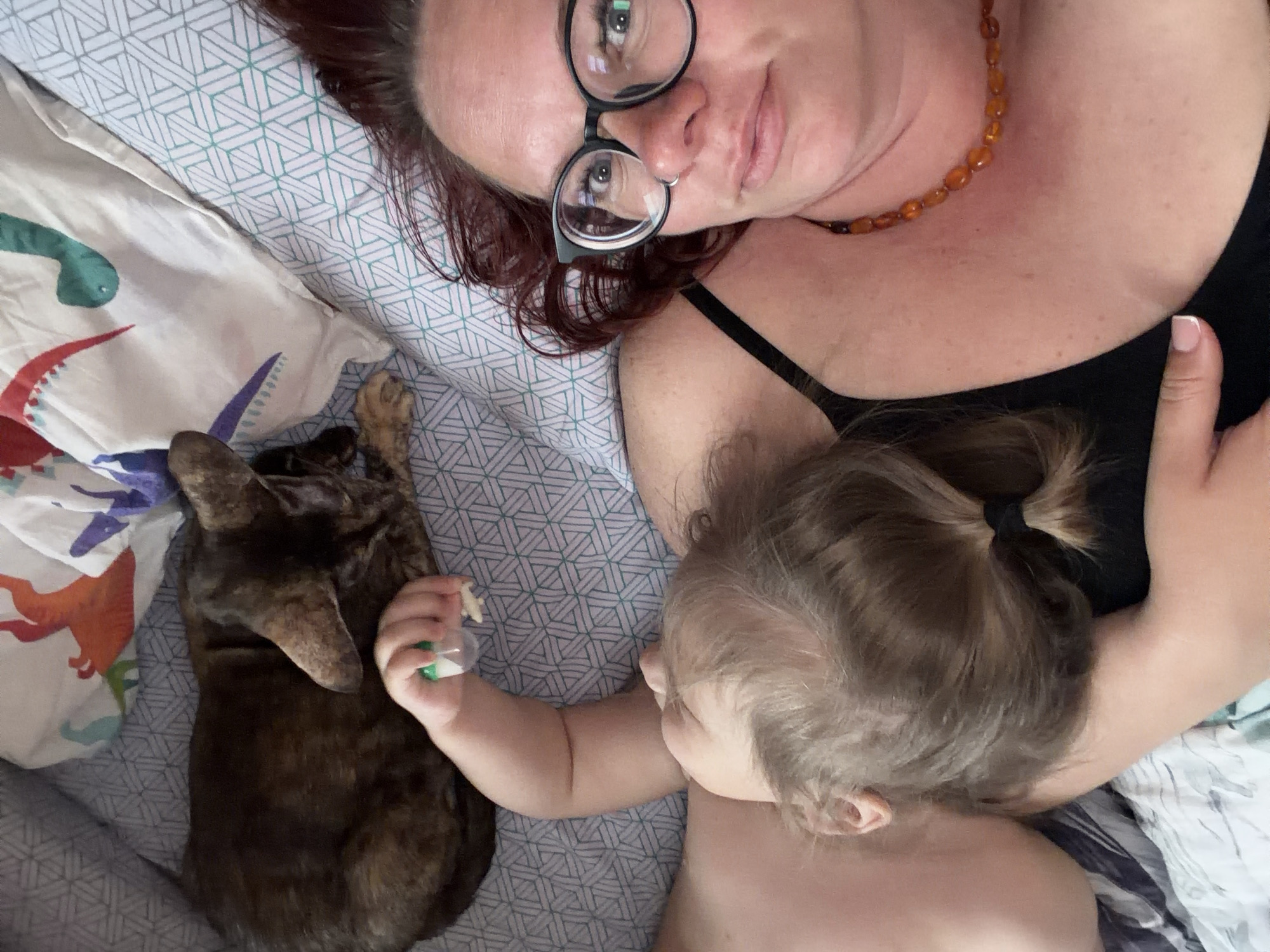 A woman with red hair and glasses is lying on a patterned bedsheet with a dinosaur-themed pillow, alongside a toddler and a brown dog. The toddler is holding a small toy in their hand. The woman is wearing a black top and a necklace. The location is possibly a domestic setting, but not definitively identified as Chaikovsky, Russia.