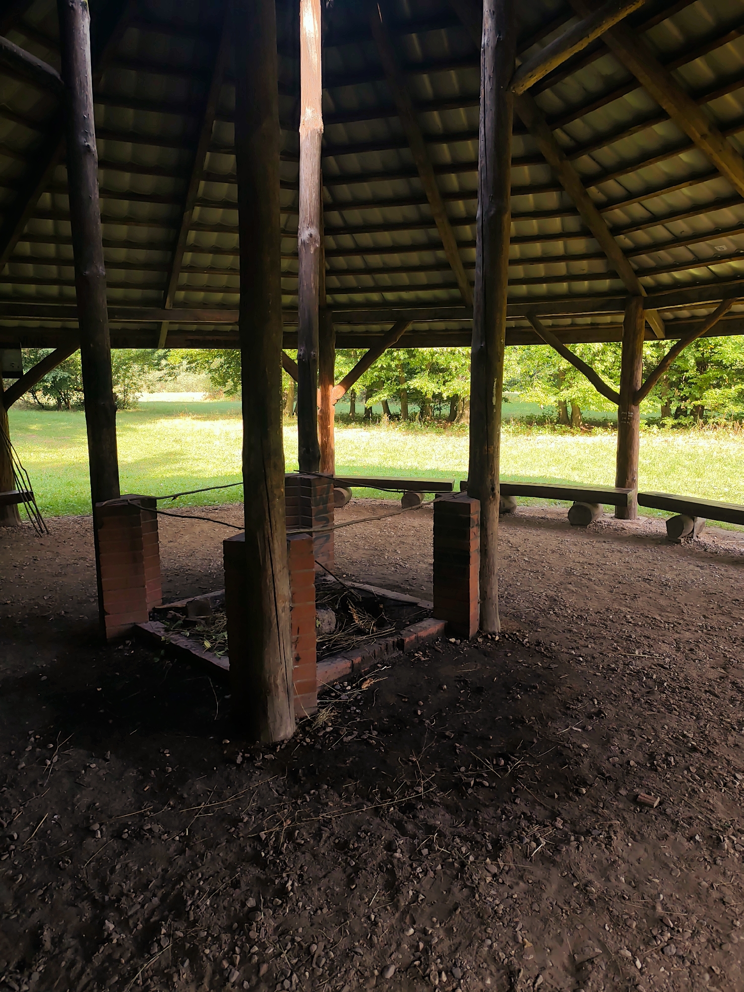 This image shows the interior of a rustic outdoor pavilion with a thatched roof supported by wooden posts. In the center of the pavilion is a brick fire pit with remnants of wood and debris. Several wooden benches are situated around the perimeter of the pavilion, facing outwards towards a grassy area with trees in the background. The ground inside the pavilion is covered in dirt and small stones. The scene appears to be set in a natural, wooded environment.