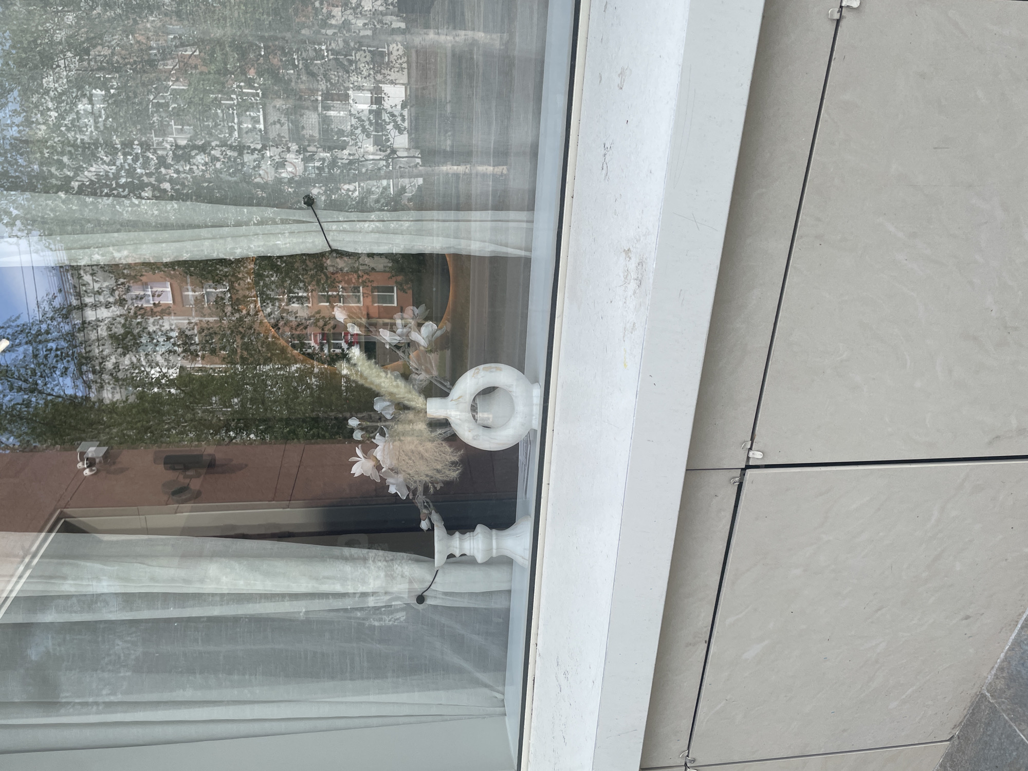 The image depicts a reflection in a window, likely in Surgut, Russia, given the context of the user's request. The reflection shows a multi-story building with a brick facade and numerous windows, along with trees. Inside the window, on a white ledge, are two decorative objects: a white candlestick on the left and a white, circular vase with an opening in the center, containing a small bouquet of dried flowers and artificial white blossoms, on the right. White sheer curtains are visible, partially obscuring the view through the window. The exterior of the window ledge is made of light-colored stone tiles.