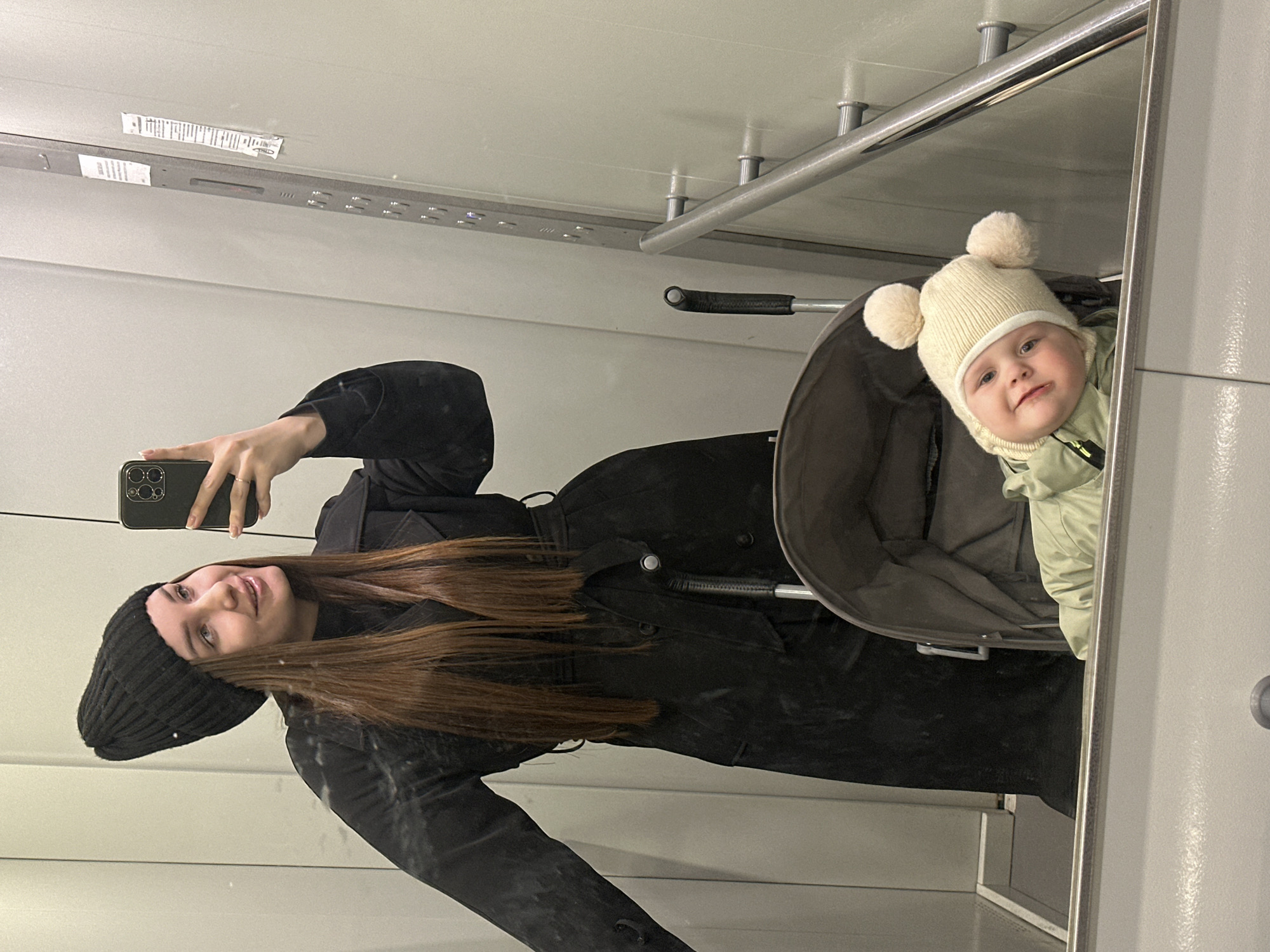 A woman with long brown hair wearing a black beanie and black coat is taking a selfie in a mirror with a baby wearing a white hat with pom-poms. The baby is sitting in a stroller. The reflection suggests that the scene is indoors, with a simple, utilitarian interior. The location cannot be determined from this image alone, nor if it is in Chaikovsky, Russia.