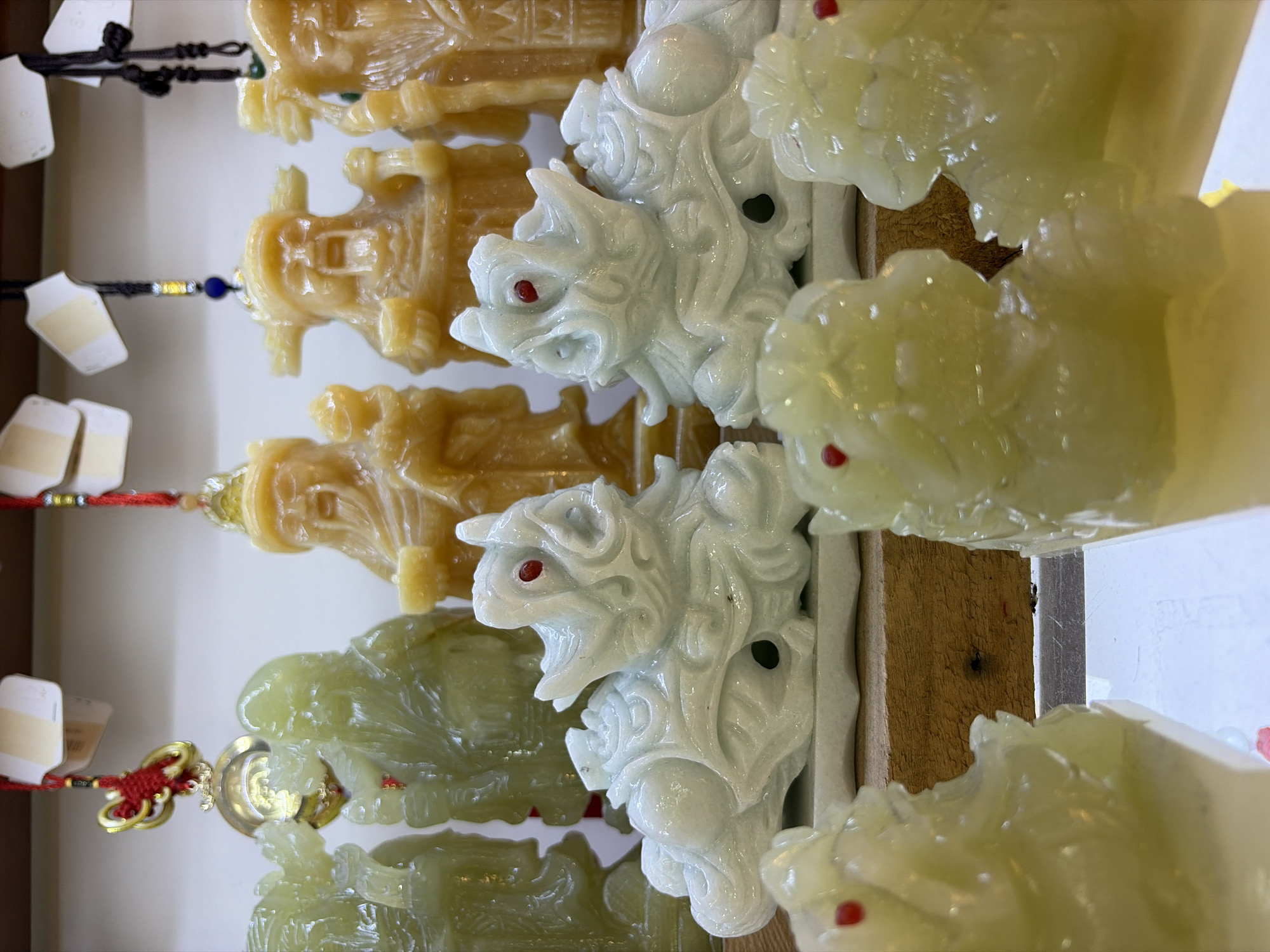 A shelf in what appears to be a market stall in Da Nang, Vietnam, displays several jade-like carved figurines. In the foreground, a row of lighter green, translucent figures with red dot eyes are arranged. Behind them, two larger, white, translucent carved mythical creatures, possibly foo dogs or dragons, with red dot eyes are positioned. In the background, several larger, opaque, golden-yellow carved figurines of men with beards and elaborate hats stand in a line, some with red thread tassels hanging from them. The background is blurred, suggesting the focus is on the displayed items. The figurines are arranged on shelves with wooden accents.
