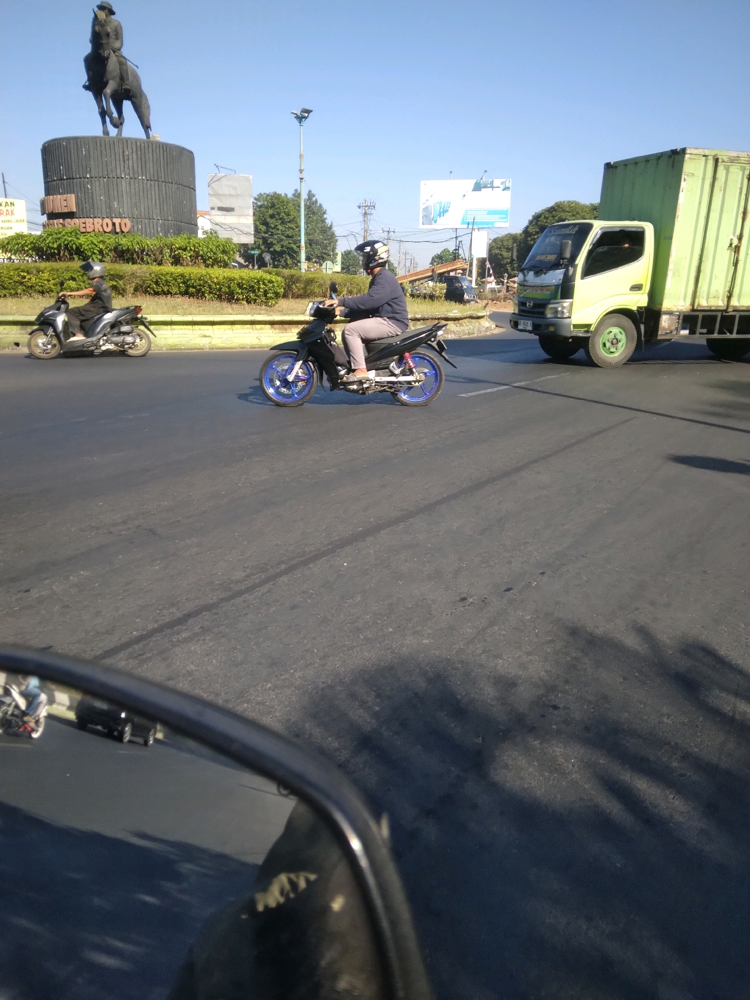The image captures a street scene in Sokaraja, Indonesia, likely a busy intersection or roundabout given the presence of a large monument. Dominating the background is a dark statue of a person on horseback, atop a cylindrical pedestal. The text "SOEBROTO" is visible on the pedestal. A bright green truck with a visible license plate "AA-9049-NB" is in the middle ground, to the right of the frame, traveling on the road. In the foreground and mid-ground, two motorcyclists are riding. The motorcyclist on the left is on a silver scooter, and the motorcyclist on the right is on a black motorcycle with blue rims, wearing a dark jacket and grey pants. The road surface is asphalt, with clear lane markings. The sky is clear and blue. A car's side mirror is visible in the bottom left corner, reflecting a portion of the road and other vehicles. Shadows of trees and possibly overhead structures are cast on the road, indicating the time of day is likely late morning or early afternoon. The environment appears to be urban, with some greenery and utility poles visible.