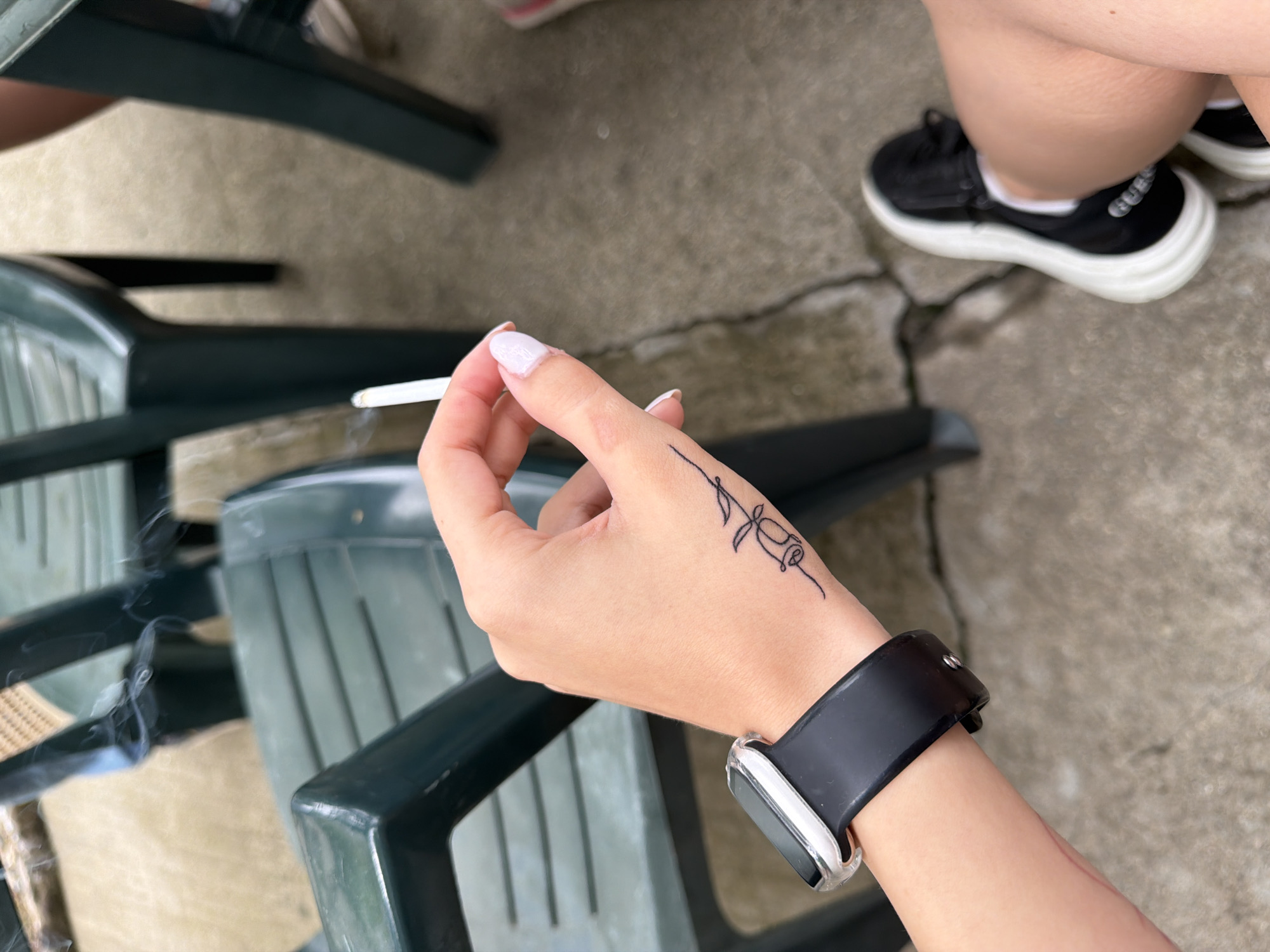 A close-up, high-angle outdoor shot captures a person's right hand holding a lit cigarette, from which faint wisps of smoke rise. The hand exhibits light-colored nail polish on the fingernails and a small, stylized black line-art tattoo resembling a flower or rose on the back of the thumb-side. A smartwatch with a clear protective casing and a black silicone band is worn on the wrist, with a thin red linear mark visible on the inner forearm below the watch. The scene's background features a concrete ground with visible cracks and several dark green plastic chairs, indicating a casual outdoor setting such as a patio. Part of a bare leg and a black sneaker with a thick white sole are visible in the lower right corner.