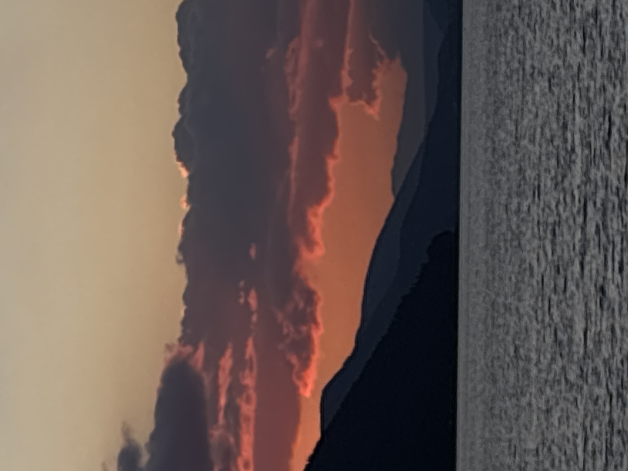 The image depicts a tranquil body of water in the foreground, reflecting the muted tones of a dusky sky. In the middle ground, a range of dark, silhouetted mountains forms a dramatic backdrop. The sky above is a canvas of warm hues, transitioning from soft peach and orange near the horizon to a pale yellow higher up. Wispy clouds, tinged with vibrant pinks and purples from the setting sun, are scattered across the upper portion of the scene, adding depth and texture. The overall impression is one of serene natural beauty at the close of day.