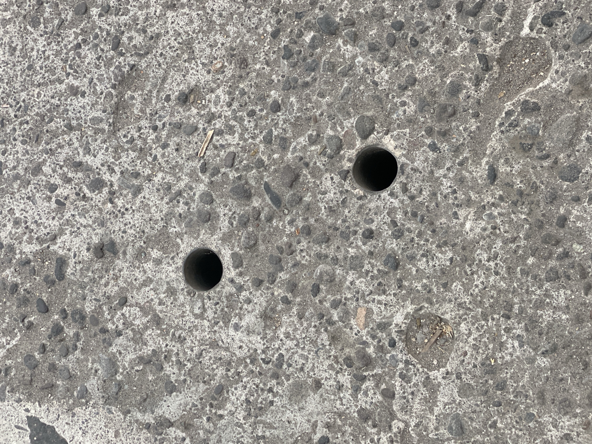 The image shows a close-up, overhead view of a concrete surface with embedded stones and debris. Two circular holes, appearing to be drains or openings, are visible in the concrete. One hole is in the upper left quadrant of the image, and another is in the lower right quadrant. A small pile of dirt and debris, including a thin twig, is present in the lower left quadrant, near the edge of the frame. The concrete appears weathered and uneven. The location is identified as Kasihan, Indonesia.