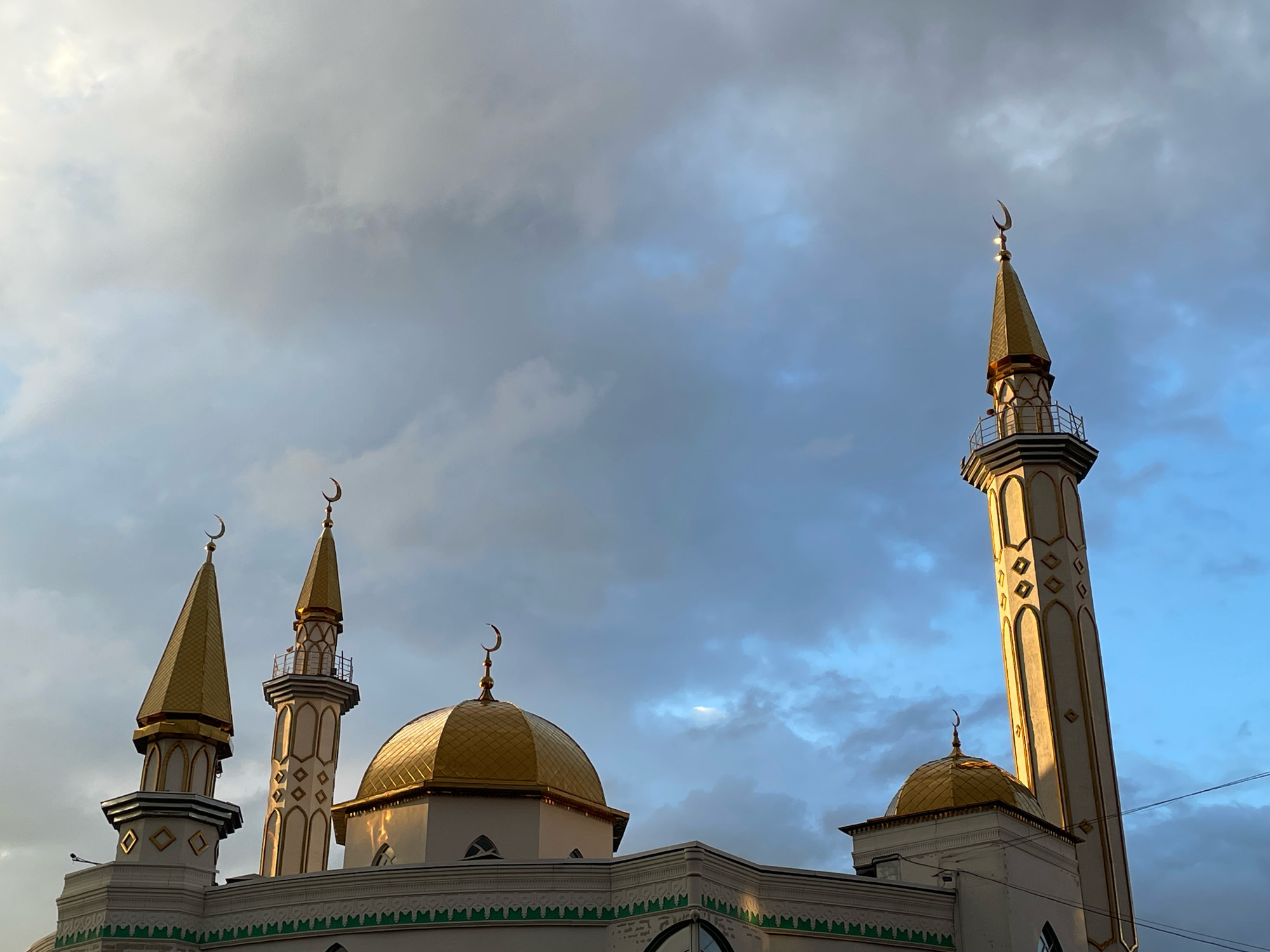 The image shows the mosque in Izhevsk, Russia, under a cloudy sky. The mosque features multiple golden domes and tall minarets topped with crescent moons. The architecture is predominantly white with gold accents on the domes and minaret details. The sky transitions from a lighter, overcast gray on the left to a clearer blue on the right, with streaks of sunlight breaking through the clouds and illuminating parts of the minarets. There are no people visible in the frame. The environment appears to be urban, with a hint of power lines in the far right of the image.