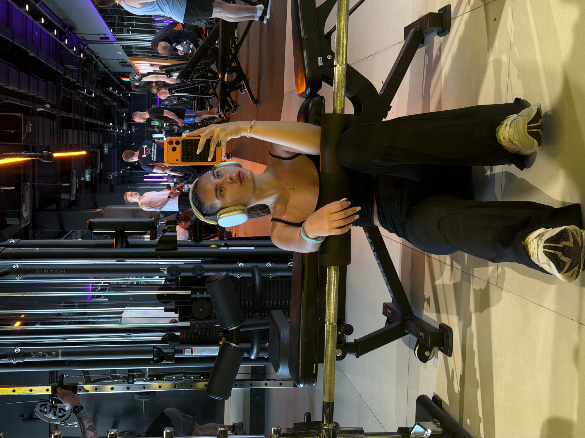 The image depicts a modern gymnasium located in Krasnoyarsk, Russia. In the foreground, a young female with dark hair pulled back is seated on a light-colored tiled floor, leaning against a black adjustable weight bench. She is wearing a black spaghetti strap crop top, loose black pants, and light-colored athletic shoes with dark soles. She has light-colored over-ear headphones on, a gold cross pendant necklace, a silver bracelet on her right wrist, and a light blue bracelet on her left wrist. She holds an orange-cased smartphone with her right hand, appearing to take a mirror selfie, with her gaze directed towards the phone/mirror. Her left arm rests on her left leg.
Behind her, the gym environment is active. Various weightlifting and exercise machines are visible. A prominent multi-station cable machine with numerous vertical cables and weight stacks stands directly behind the female, bearing a mirror-reversed "MATRIX" brand logo. To the far left, a man with tattoos on his left arm is operating a piece of equipment, possibly adjusting weights on a smith machine or cable system. Further into the midground, two men stand conversing; one wears a white t-shirt, and the other a blue t-shirt with a logo. To the right, another man is performing an incline dumbbell press on a black adjustable bench, while a partially visible man in a light blue tank top stands further right.
The gym features dark walls and large mirrored sections reflecting the interior. The ceiling is industrial-style with exposed ducts, pipes, and linear light fixtures. Overhead lighting includes warm orange-toned strip lights and cooler purple/blue accent lighting, particularly near the mirrored areas. Green exit signs are mounted overhead. The floor throughout the gym is a consistent light-colored tile.