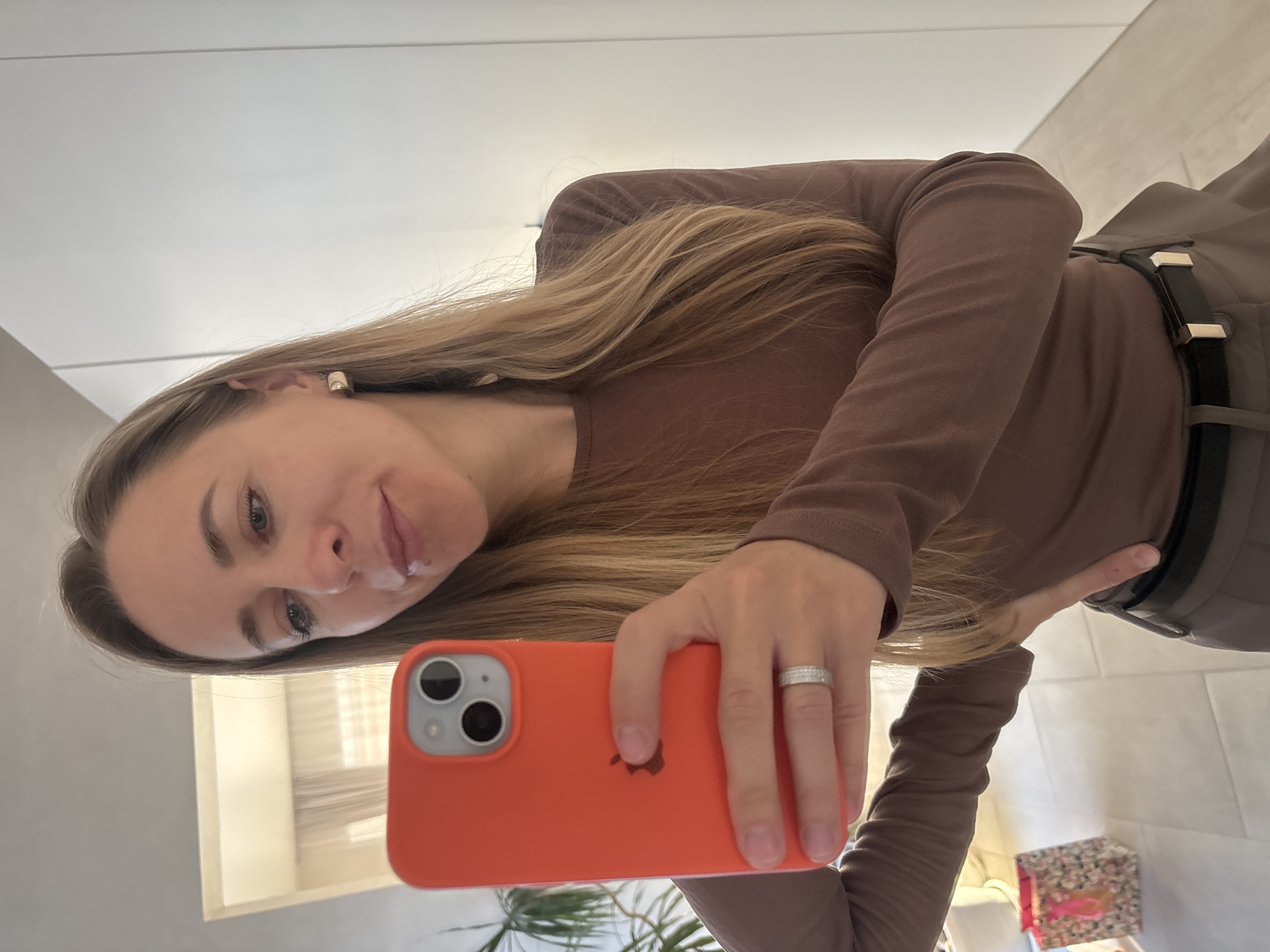A woman with long blonde hair is taking a selfie in what appears to be an indoor environment. She is wearing a brown, long-sleeved shirt. The phone she is using has a bright orange case. A ring is visible on her ring finger. In the background, there is a glimpse of plants, white walls, and what seems to be furniture or other objects. The location is Chaikovsky, Russia.