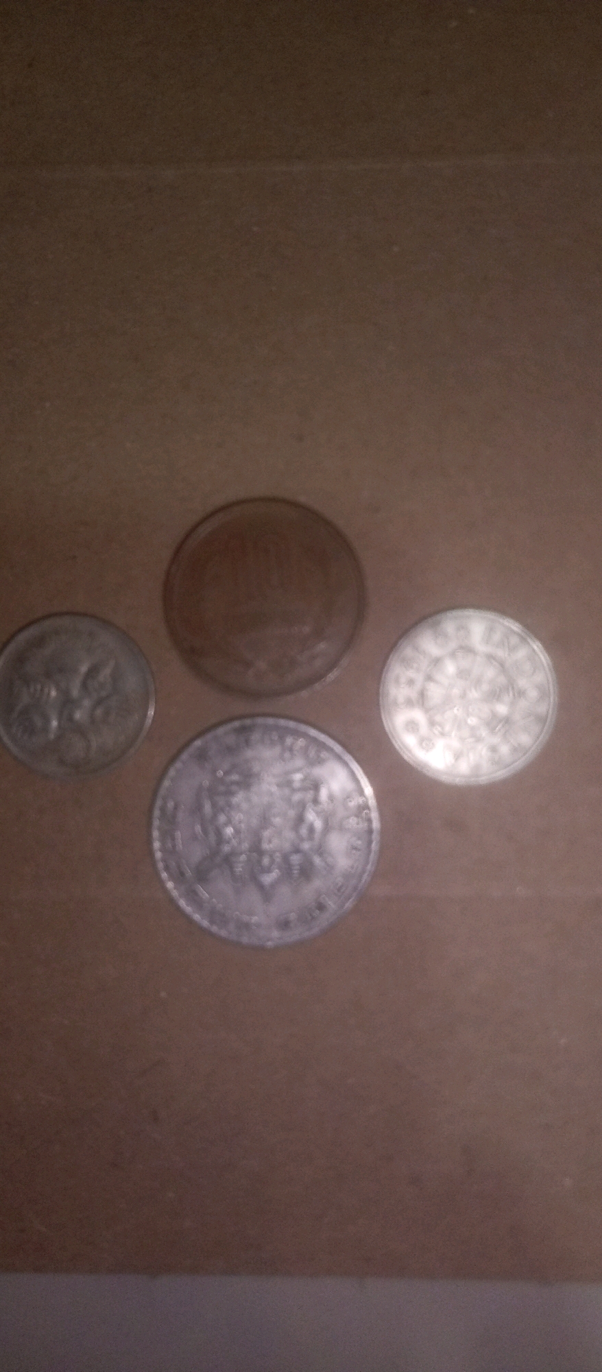 The image shows four coins arranged on a brown surface, possibly cardboard. The coins are of different sizes and appear to be of varying metallic compositions. One coin, located in the upper center, is darker and appears to be made of a coppery material, with some indistinct markings. To its left, a smaller, lighter-colored coin is visible, displaying an intricate design. Below and to the right of the coppery coin is another lighter-colored coin with a detailed pattern around its edge and a central design. The largest coin, positioned in the lower center, is a lighter silver color and features prominent lettering and what appears to be a depiction of multiple figures. The lighting is dim, casting faint shadows. The background is a plain, textured brown surface. There is no discernible connection to the city of Kediri City, Indonesia, as there are no contextual elements in the image to suggest this location.