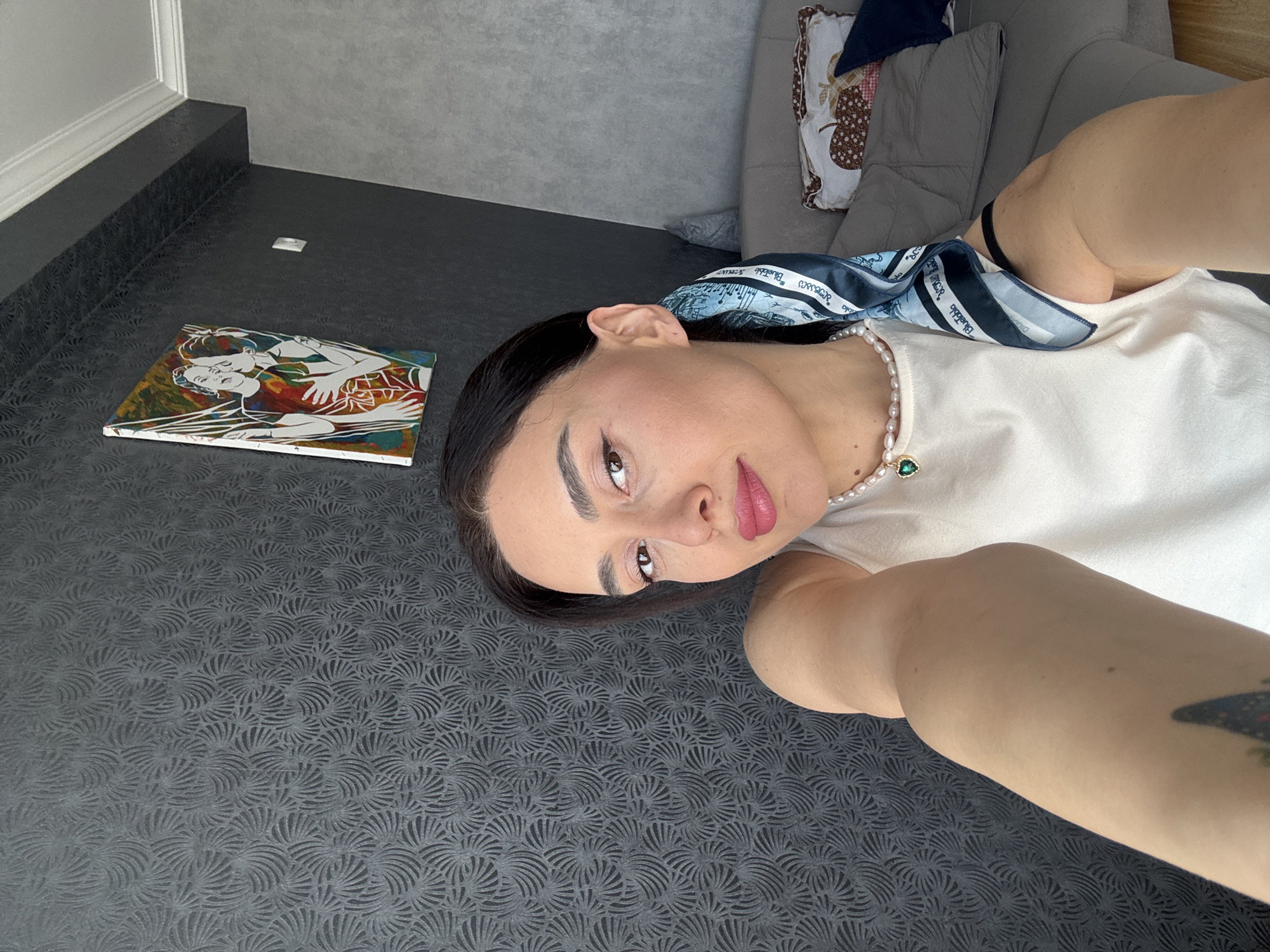 The image captures a close-up selfie of a woman lying down in an indoor setting. She has dark hair, defined eyebrows, winged eyeliner, and pink lipstick. She wears a white top, a pearl necklace with a green emerald-like pendant, and a light blue patterned scarf around her neck. A colorful tattoo, appearing to be a butterfly or insect, is partially visible on her right forearm. Her expression is calm as she looks towards the camera.
The background features a dark grey or black floor covered in a repeating textured pattern of stylized fan or scallop shapes. A dark wall or baseboard with a subtle texture and a white trim is visible along the edge. In the upper right, a grey sofa with a patterned cushion (featuring brown and white designs) can be seen. On the floor, next to the woman, lies a rectangular piece of artwork. The painting depicts two stylized figures, possibly embracing, with white outlines against a vibrant, abstract background of red, blue, green, and yellow hues.
The scarf worn by the woman displays visible text, including "Bluebottle" and the Georgian script "საქართველო" (Sakartvelo), which translates to "Georgia." This directly correlates with the provided location context of Tbilisi, Georgia. The lighting suggests it is daytime.