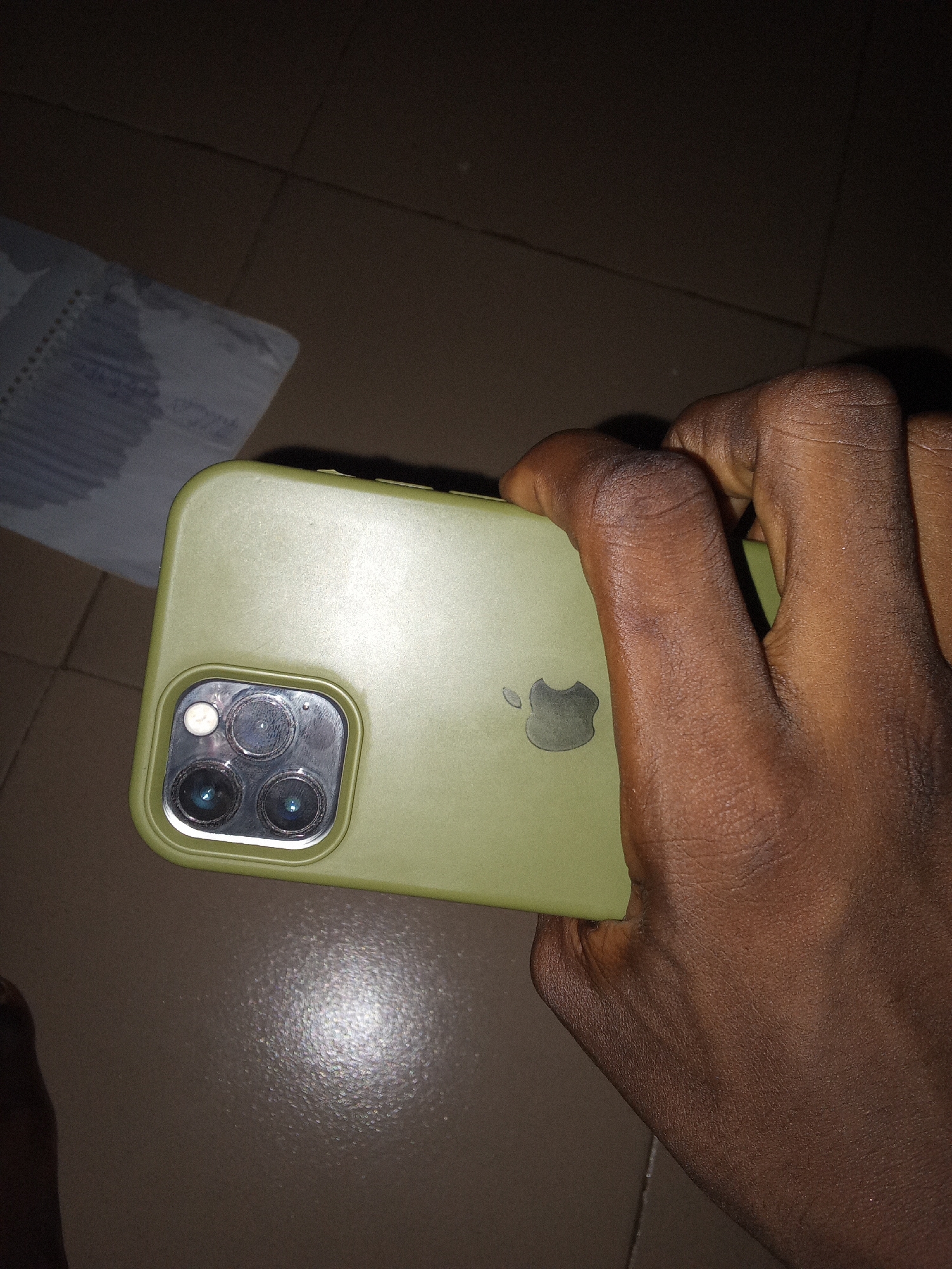 A dark-skinned hand is holding a light green iPhone 11 Pro in Safari, Nigeria. The phone is oriented horizontally with the cameras facing the viewer. The camera module, containing three lenses and a flash, is visible on the left side of the phone. The Apple logo is embossed on the back of the phone. The background consists of a tiled floor.