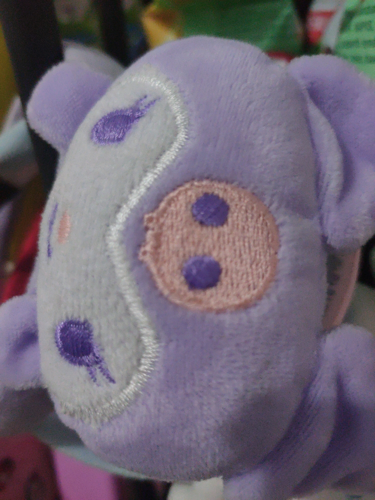 A close-up, low-angle, indoor photograph captures a purple plush toy with white and pink embroidered facial features. The toy, likely a stuffed animal, has large, light grey circular markings on its face, each with a smaller purple embroidered detail. A pink embroidered oval shape with two purple circular elements is also visible on the toy's face. The background is blurred and shows hints of other objects and possibly clothing. The toy is positioned centrally in the frame, and its texture appears soft and fuzzy. The image's composition focuses on the toy's head and upper body. The lighting suggests an indoor environment, possibly a room with soft, diffused light. There is no discernible connection to Batam, Indonesia.