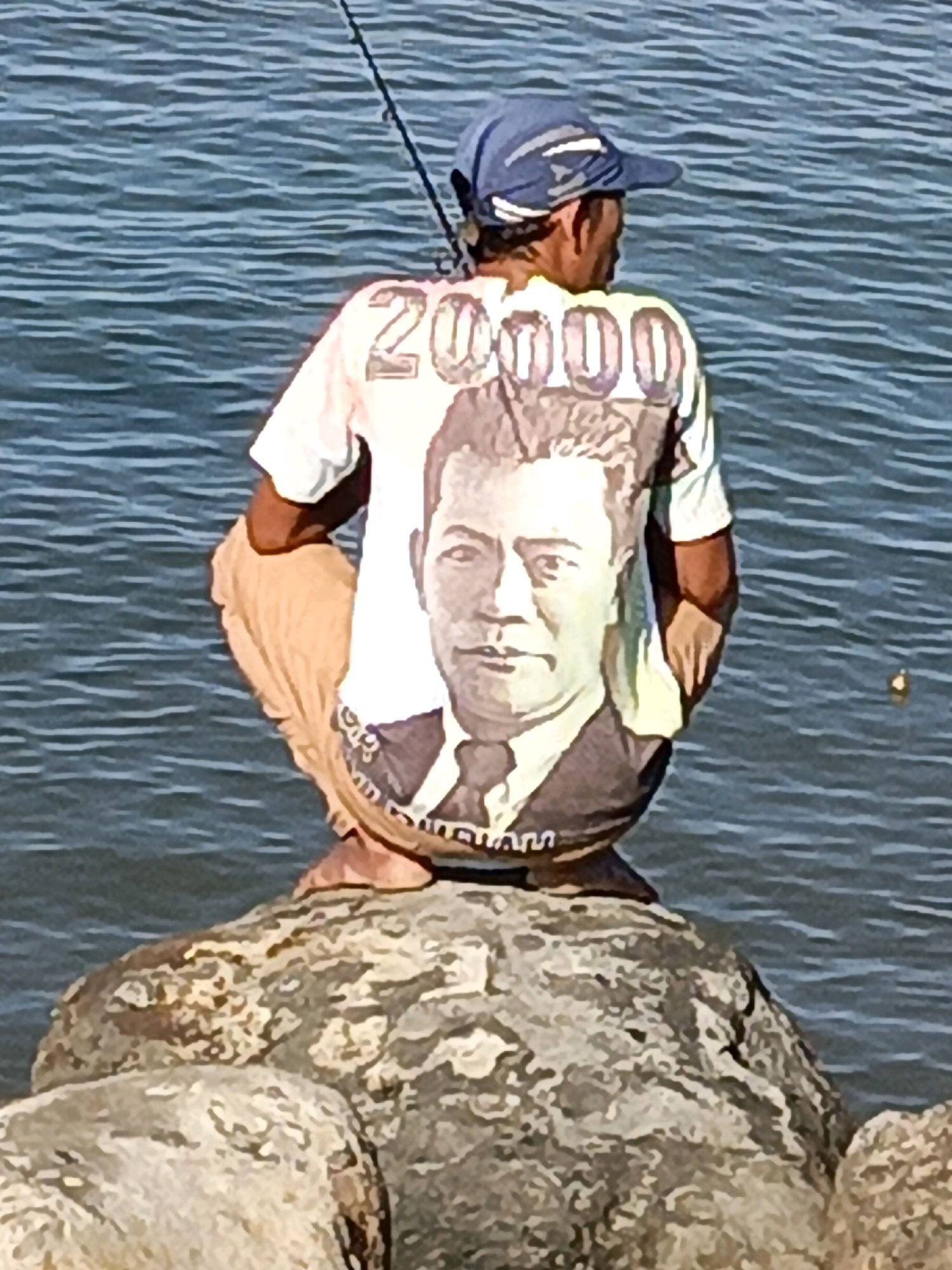 A man fishing on a rocky outcrop beside the sea in Bulukumba, Indonesia. The man is wearing a blue baseball cap and a white t-shirt with a large image of a man's face and the numbers "20000" printed on the back. He is wearing light-colored shorts. The water is blue and slightly choppy, with a fishing rod extending into the frame from the top. The man is crouched down on the rocks, facing away from the camera.