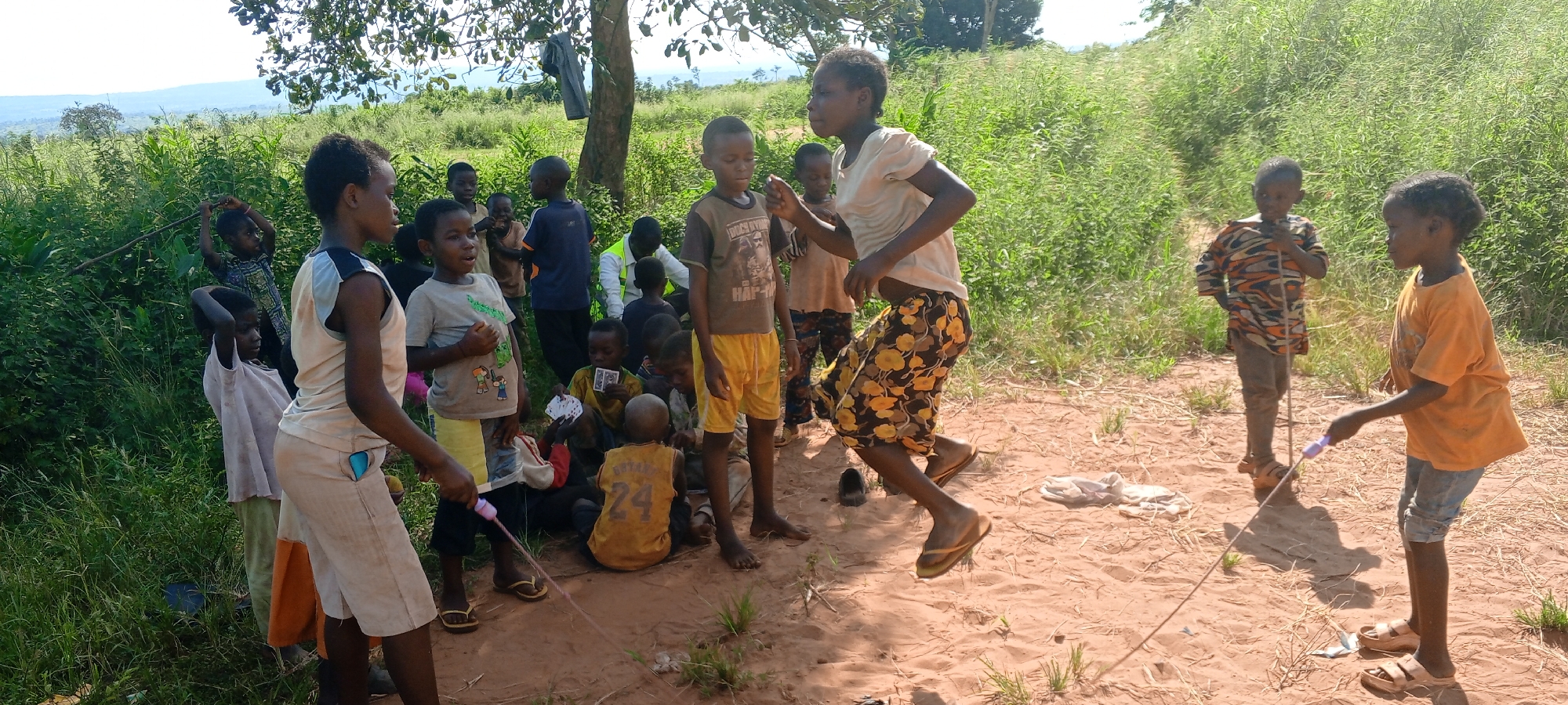 The image depicts a group of approximately 20 children in what appears to be a rural setting near Ngalula, Democratic Republic of the Congo. The ground is sandy and sparsely covered with grass, surrounded by dense green vegetation. Several children are grouped together, some sitting on the ground playing cards, while others stand and watch. Two children are actively engaged in a jump rope game. Other children are standing at the edge of the group looking on. The clothing worn by the children is varied and casual, and many are barefoot or wear simple sandals. In the background, a tree provides some shade, and there is a glimpse of rolling hills or mountains in the distance.