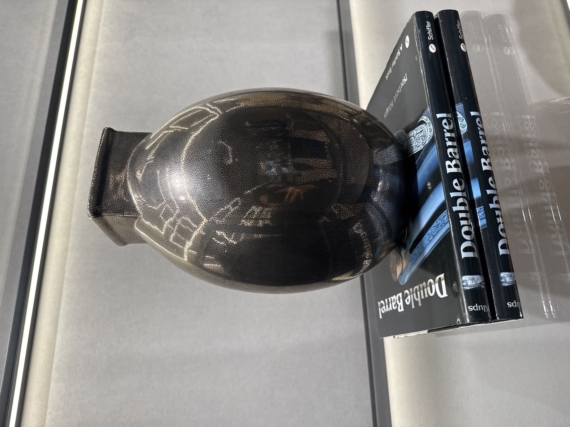 The image presents a still life composition. A black, textured football with silver markings is positioned on a gray surface. Behind the football, three copies of a book titled "Double Barrel" by Norbert Klups, published by Schiffer, are stacked vertically. The spine of each book is visible. The setting is indoors, as indicated by the presence of artificial lighting elements. The image does not contain any information to determine if the location is the city of Dubai, United Arab Emirates.