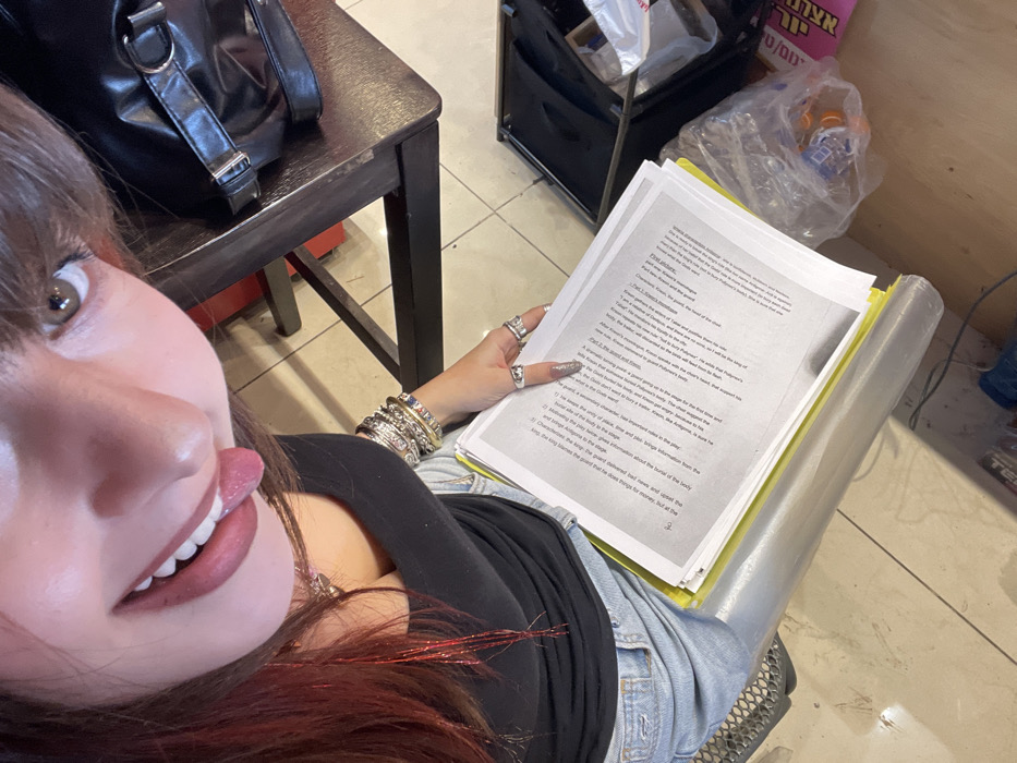 An image depicts a female individual partially visible from a high-angle, downward perspective within an interior location in Raanana, Israel. The individual has brown hair with red highlights and bangs, and is looking upward towards the camera with her mouth open, tongue extended. She is wearing a black top and light-wash denim jeans, a silver-colored pendant necklace, multiple silver-colored bangles on her left wrist, and several silver-colored rings on her right hand. Her fingernails are long and manicured. She is holding a thick stack of white papers with printed text, resting on a yellow folder, on her lap. The immediate environment includes a light-colored tiled floor. To the right, a dark-colored table or counter surface is visible, upon which rests a black bag with a silver-colored buckle. Below the table, an open black case containing various items is present, along with a clear plastic bag holding orange objects and a partially visible pink container with text. A rolled-up clear plastic sheet is on the floor to the left, and a dark-colored chair or stool base is partially visible on the far left.