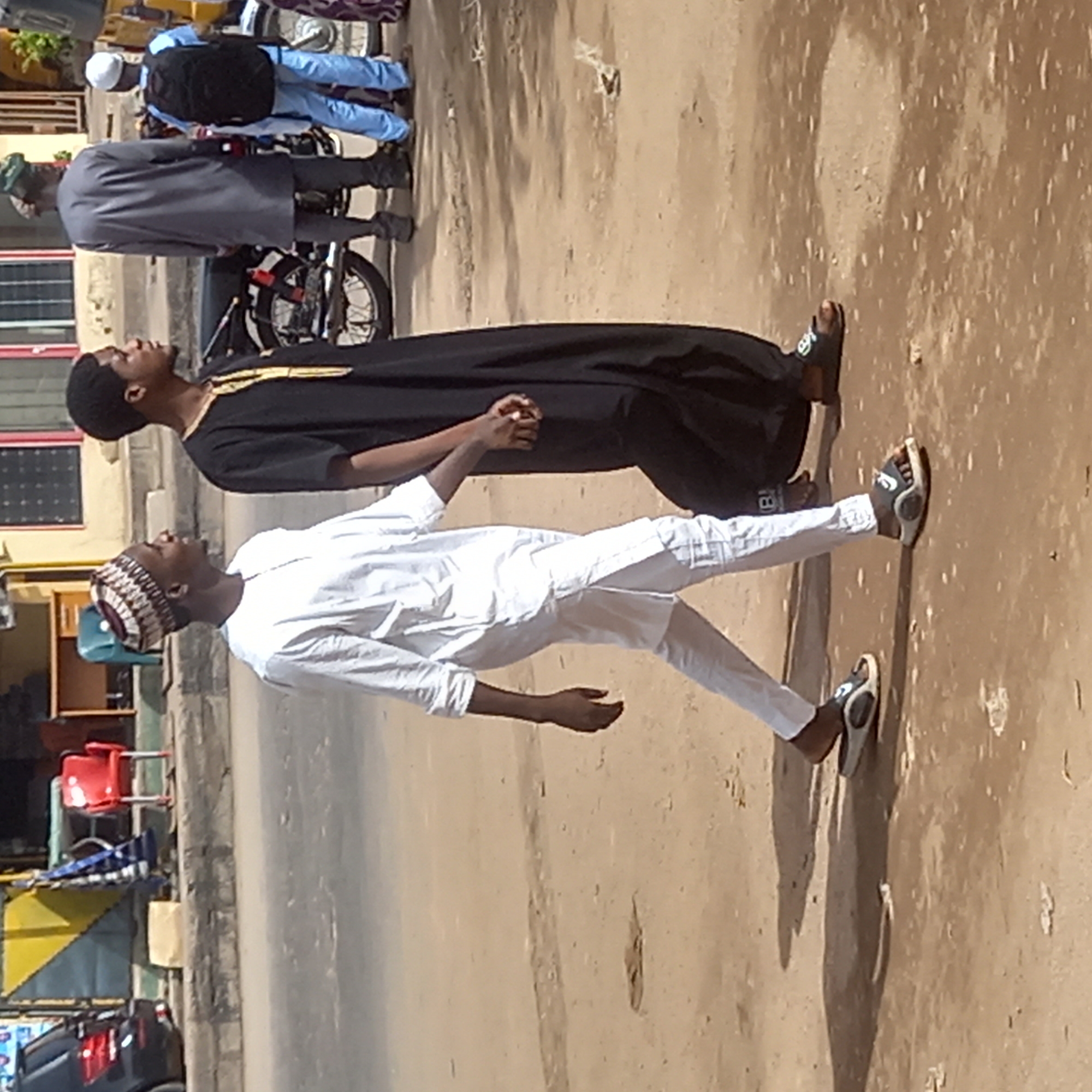 Two men are walking on a paved road, likely in Dadi Riba, Nigeria. One man is wearing a white outfit and a striped cap, while the other is in a black robe with gold trim. Their hands are clasped together. Behind them, another man in gray walks by a parked motorcycle. Further back, there's a building with windows and an open storefront with chairs and a visible portion of a car. The road has a dirt shoulder.