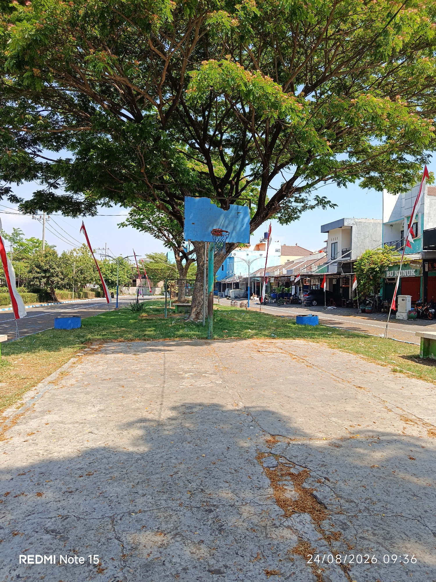 An outdoor scene in the city of Keboananom, Indonesia, features a basketball court in the foreground under a large tree. The court surface is concrete and shows some cracks and debris. A blue basketball hoop with a red rim and net stands prominently in the center. Several red and white Indonesian flags are visible along the street behind the court, indicating a possible national holiday or event. The background includes a street with parked motorcycles and cars, lined with a row of buildings that appear to be shops or residences. The sky is clear and blue. The timestamp "24/08/2026 09:36" and "REDMI Note 15" are visible in the bottom right and left corners of the image respectively.