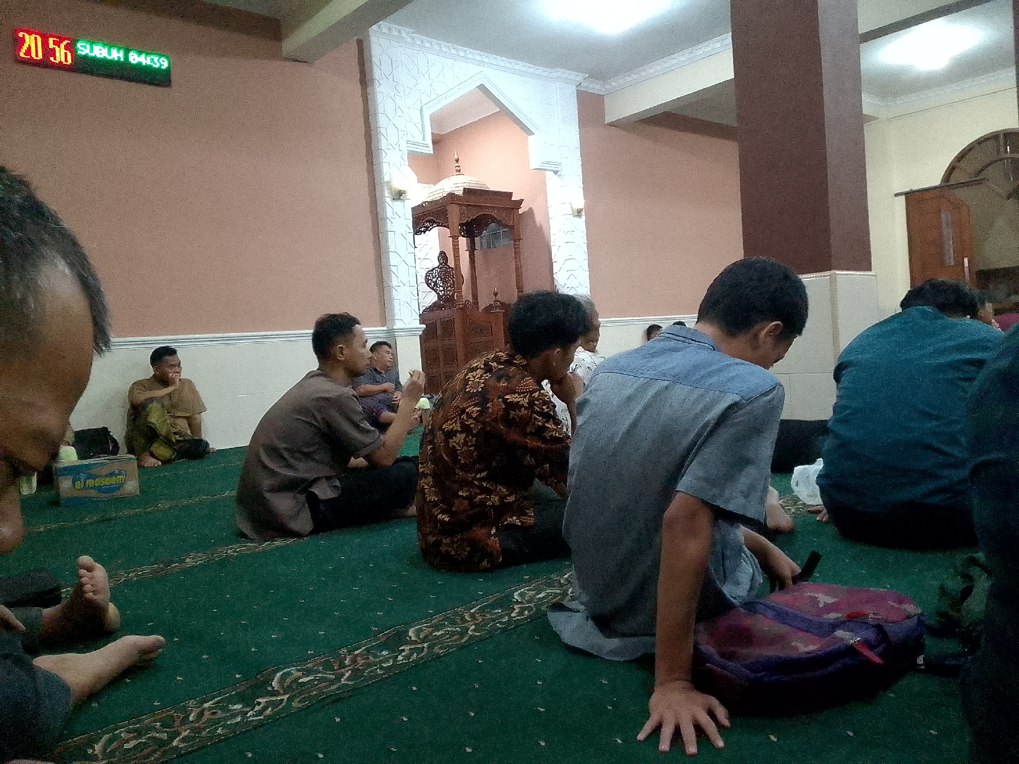 A group of men are seated on a green carpet inside a mosque. A digital clock on the wall displays "20 56 SUBUH 04:39". One man in the foreground is wearing a blue shirt and appears to be praying. Another man in a patterned brown shirt is visible behind him. To the left, a man in a brown shirt is seated with his legs crossed. A blue box labeled "al masoem" is on the floor near the front left. The mosque features ornate architectural details, including a carved wooden structure and decorative white molding on the walls. The lighting is dim, with overhead lights illuminating the ceiling.