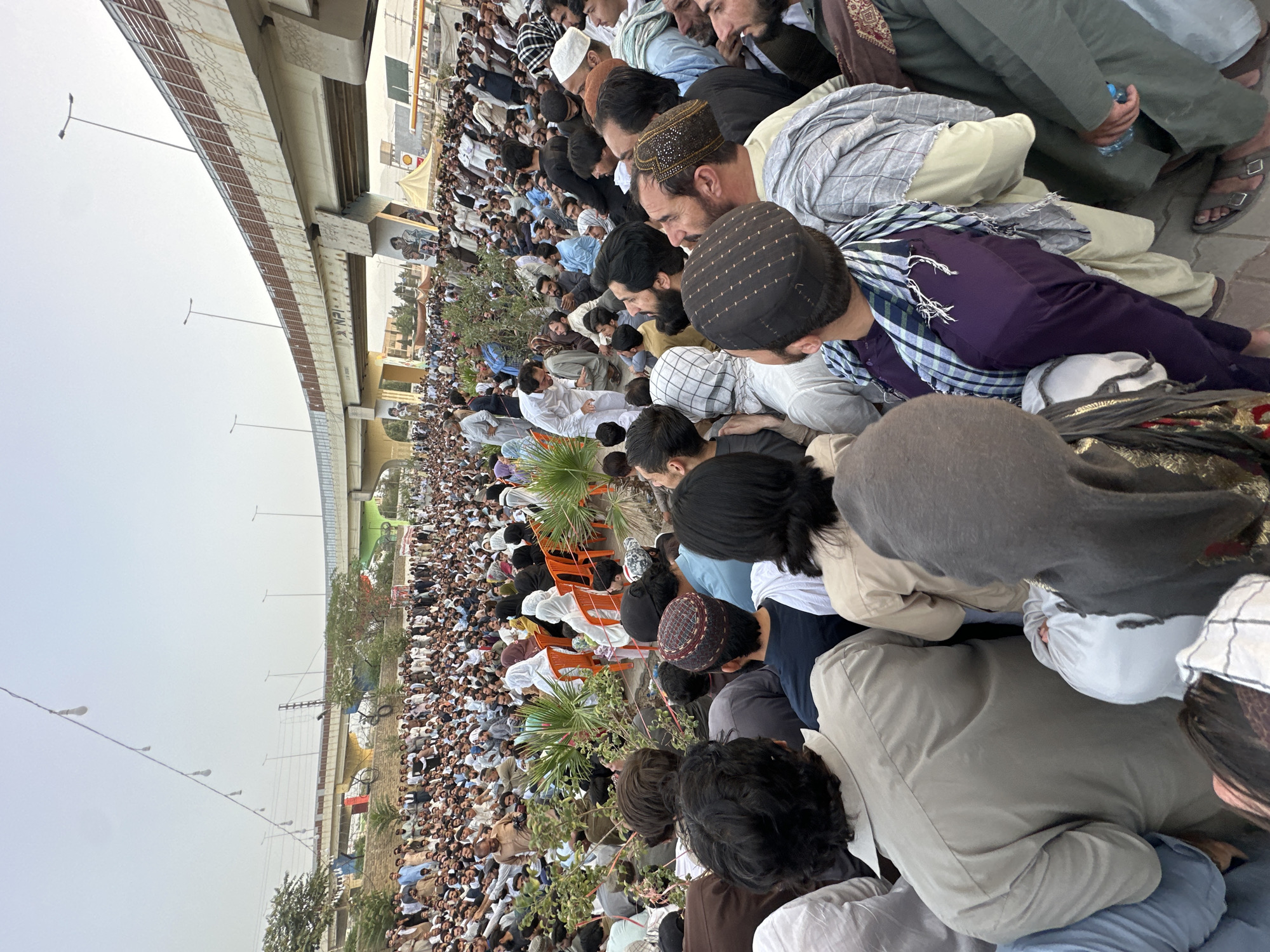 In Quetta City Tehsil, Pakistan, a large gathering of people is assembled outdoors. The scene is dominated by a crowd of individuals, predominantly men, dressed in traditional South Asian attire, including shalwar kameez and various head coverings like turbans and caps. Many are standing, while some are seated on the ground or on orange plastic chairs. Some individuals are engaged in conversation, while others appear to be listening or observing something outside the frame.
The environment includes a section of an elevated highway or flyover visible in the background, supported by concrete pillars. There are trees and foliage interspersed within the crowd and along the ground. A string of bare light bulbs hangs across the upper portion of the image, suggesting it might be evening or overcast. The ground appears to be paved or made of stone. The overall atmosphere seems to be one of a public gathering, possibly a political rally, religious event, or community assembly. The lighting suggests it is daytime, with diffused natural light.