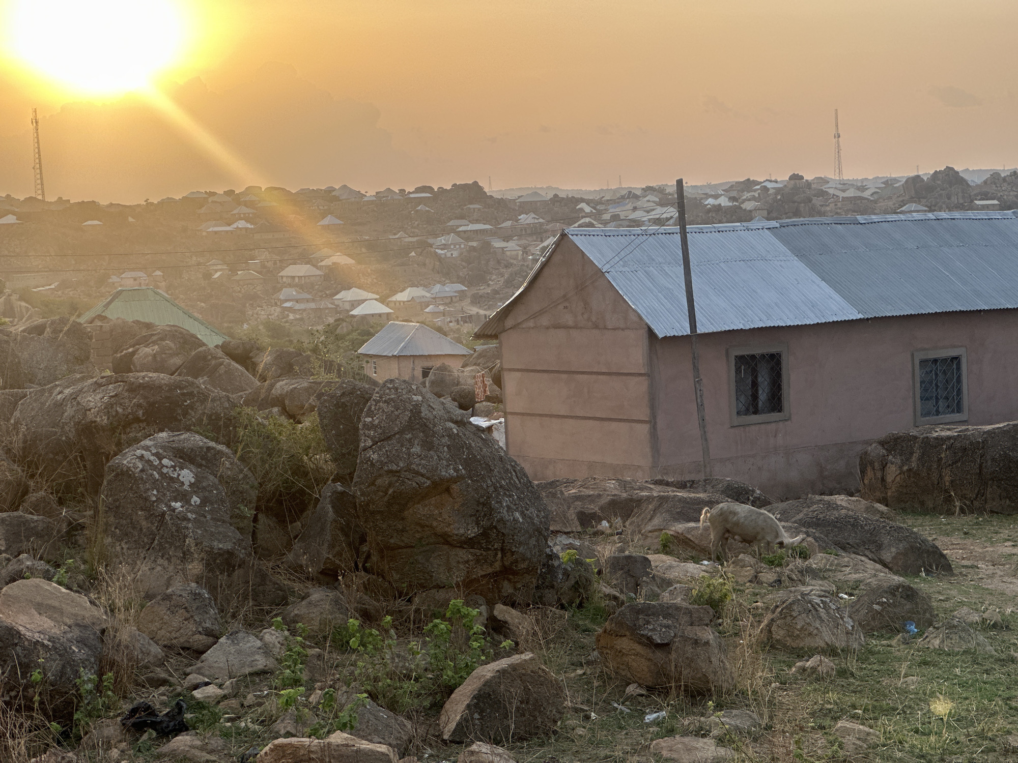The photograph shows a rural scene in Jos, Nigeria, during sunset. A small, unfinished, single-story building with a grey metal roof stands to the right of the frame. Two windows are visible on the side of the building.
The foreground is dominated by large, grey, lichen-covered rocks and dry grasses. A light-colored goat is grazing among the rocks near the building.
In the background, a cluster of buildings with light-colored roofs are seen scattered across a hilly landscape. Tall metal towers are visible on the horizon. The sky is a bright orange due to the setting sun, which is partially obscured by clouds.