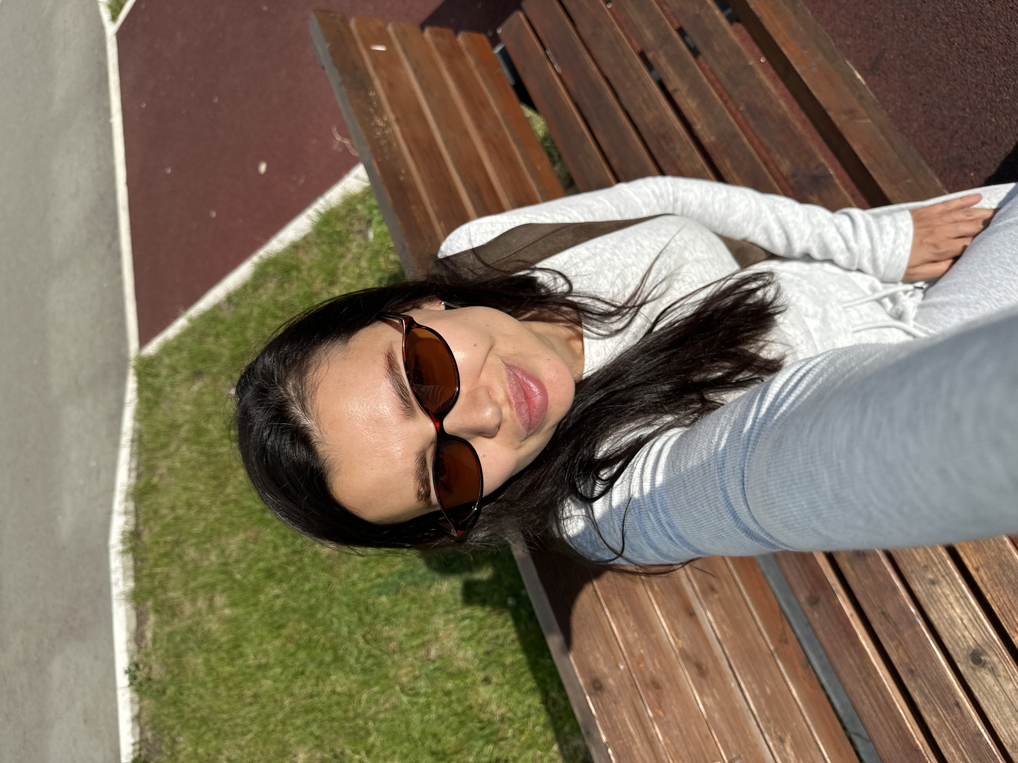 A selfie of a woman with long dark hair and sunglasses is taken outdoors. She is wearing a light gray long-sleeved top and light gray sweatpants. She is sitting on a wooden park bench. To her left is a patch of green grass and to her right is a red running track with a white border. The background consists of a gray paved surface. The lighting suggests it is daytime, possibly late afternoon due to the long shadows cast on the grass and bench. The location is identified as Chaikovsky, Russia.