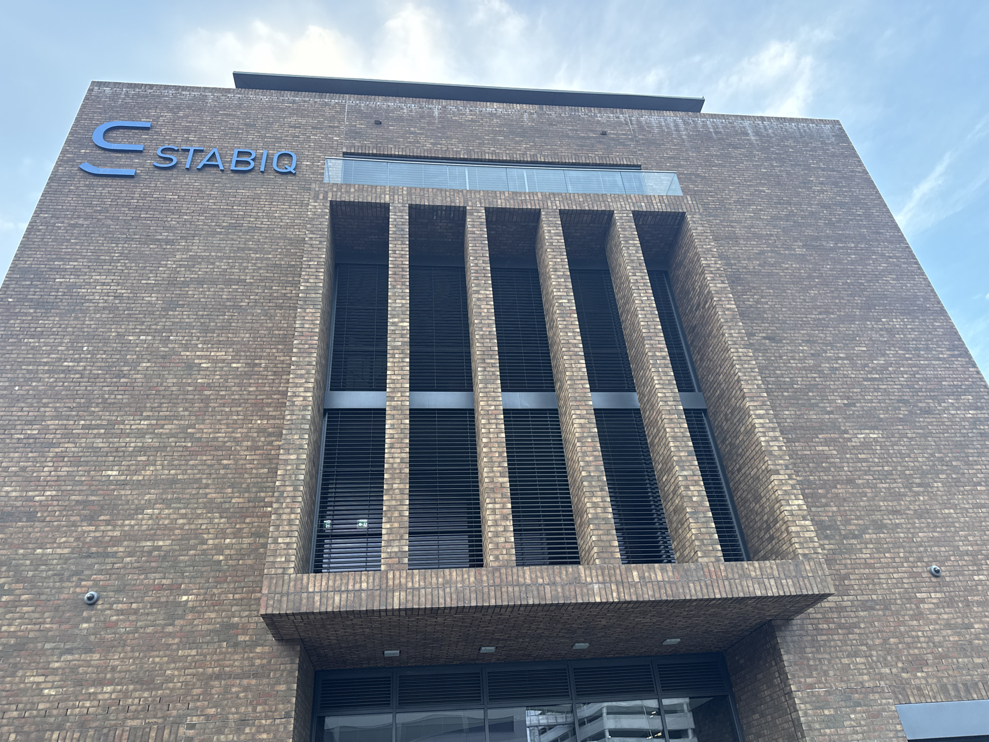 An eye-level, wide-angle shot captures the modern, brick-clad facade of a building in Nendeln, Liechtenstein. The structure features a prominent architectural detail of vertical brick columns framing dark, slatted windows. Above these, a horizontal band of glass, likely a window or balcony railing, adds a contrasting element. The "STABIQ" logo, in blue, is mounted on the upper section of the building, to the left of the vertical columns. The sky above is partially clouded, with blue sky visible between the white clouds. Security cameras are visible on the brickwork. The lower portion of the image shows a glass entrance with further slatted windows above.