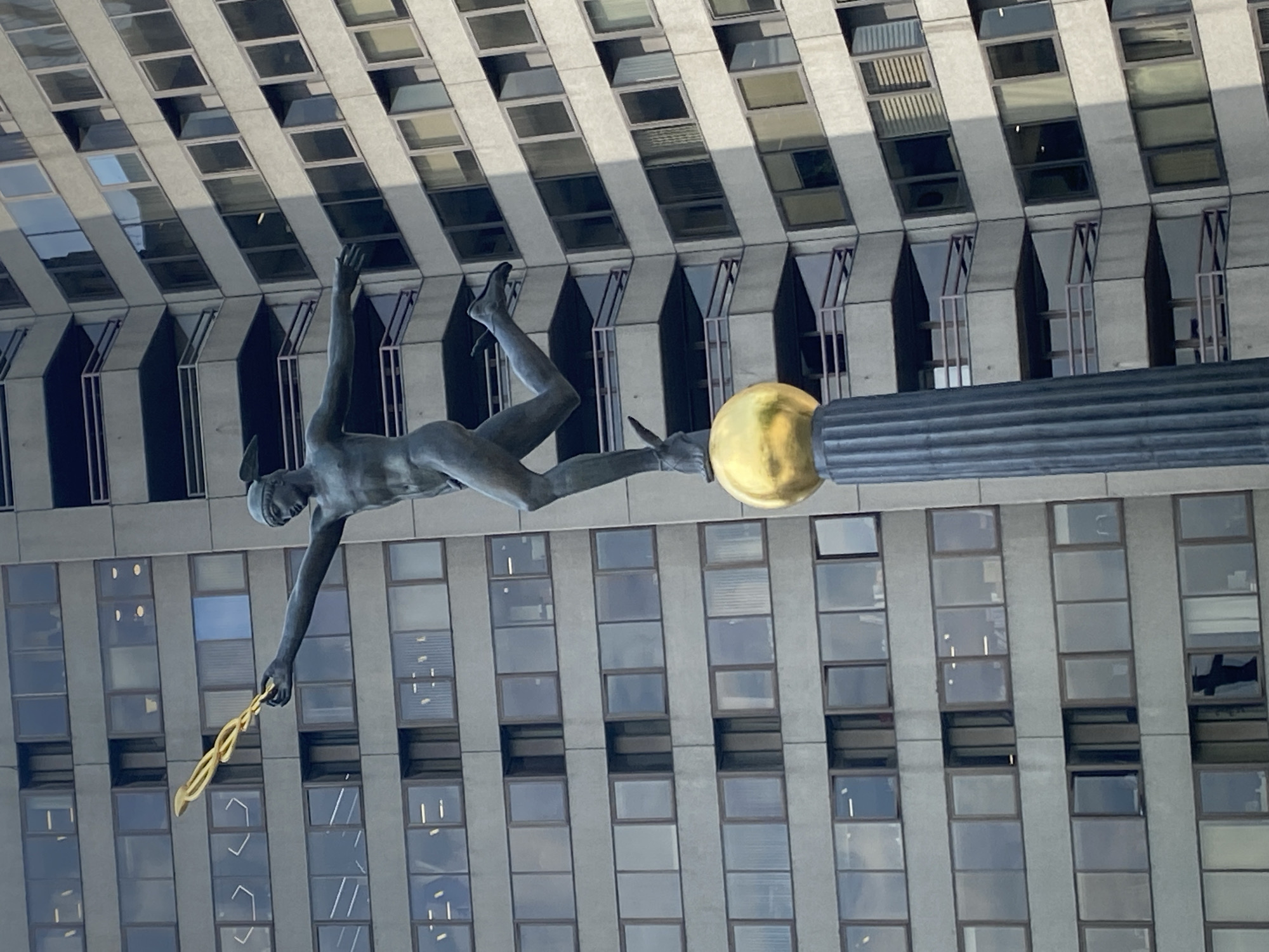 The image shows the statue of Prometheus at Rockefeller Center in New York City. The bronze statue depicts the mythological figure Prometheus lying on a gold sphere and reaching out to give fire to humanity. The backdrop is a modern skyscraper with rows of windows.
