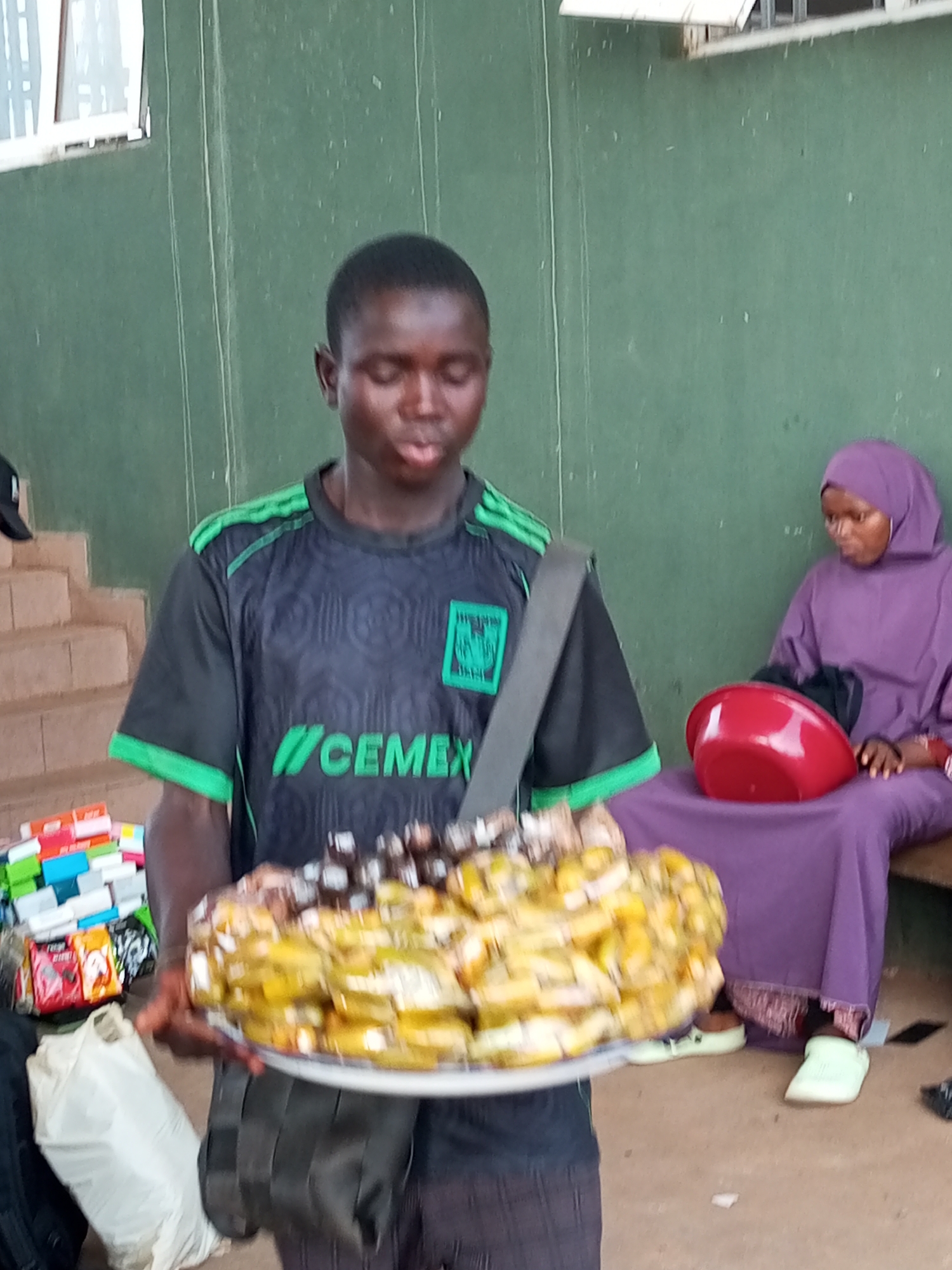 This media file depicts a young boy in a black and green t-shirt holding a large platter of what appears to be food, possibly fried plantains and some darker items. The food is arranged in a mound on a white plate. The boy is looking downwards, with his mouth slightly open. He is standing in what seems to be an outdoor setting, with a green wall behind him. To his right, a woman wearing a purple hijab and dress is seated, with a red bowl in her lap. In the background, to the boy's left, there are some stacked items, possibly goods for sale, and a set of concrete stairs. A white plastic bag is also visible in the lower left corner. The lighting suggests it could be daytime. The overall scene appears to be a marketplace or a street vendor situation. The t-shirt the boy is wearing has visible text that appears to be "CEMEX" and some green stripes on the sleeves. The woman's attire is traditional. There are no obvious cues about the time of day or weather. The emotions of the subjects are not clearly discernible.