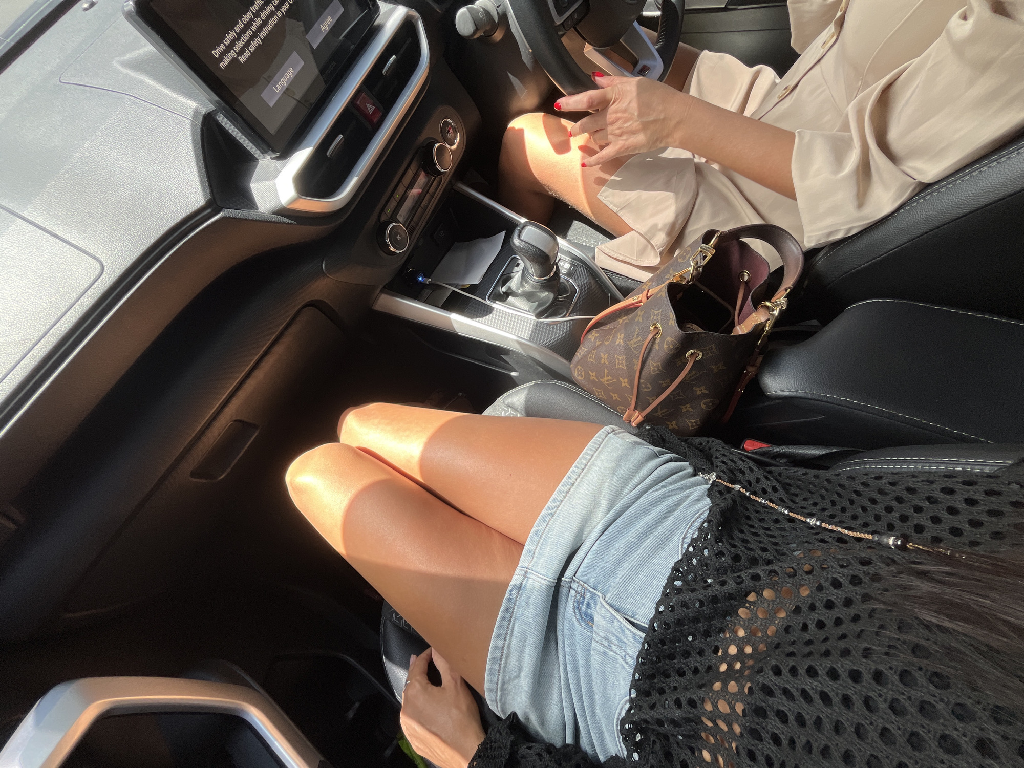 The image is taken from inside a car. Two people are visible. The driver, likely a woman, wears a tan shirt and has red nail polish. A passenger sits in the front seat, wearing denim shorts and a black mesh shirt. A Louis Vuitton handbag rests on the passenger seat. The car appears to be a newer model with a touch screen display. Based on visual information and context, the location is suspected to be Kuta Selatan, Indonesia. The environment is sunny, and the lighting casts strong shadows within the car's interior.