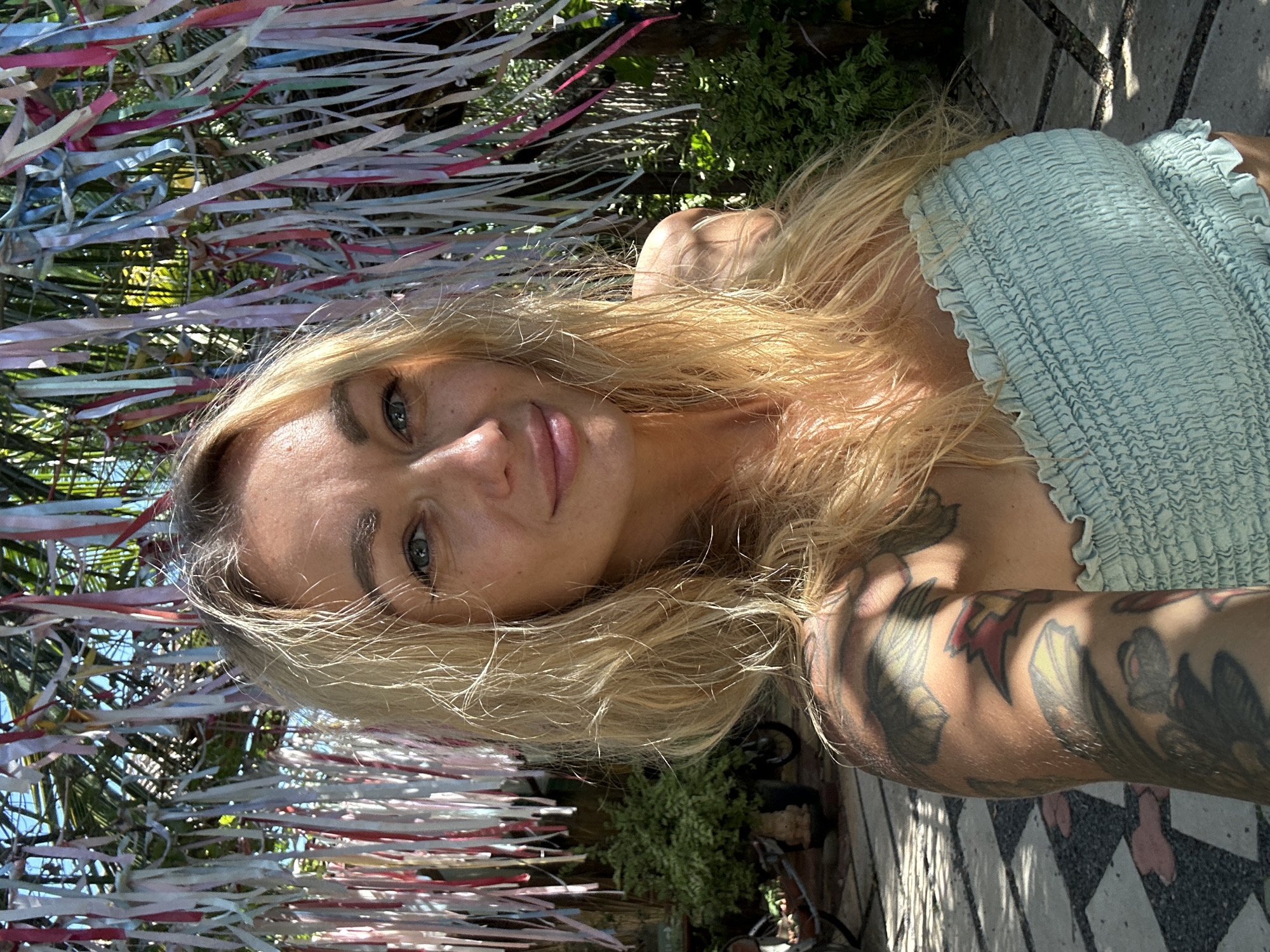 A woman with blonde hair and blue eyes takes a selfie in an outdoor setting. She is wearing a light blue smocked tube top and has tattoos on her left arm. Behind her, colorful ribbons hang from trees, and foliage and trees are visible. The ground appears to be paved with stone tiles. In the background, a motorcycle and plants in pots can be seen. The scene suggests a tropical or subtropical location.
