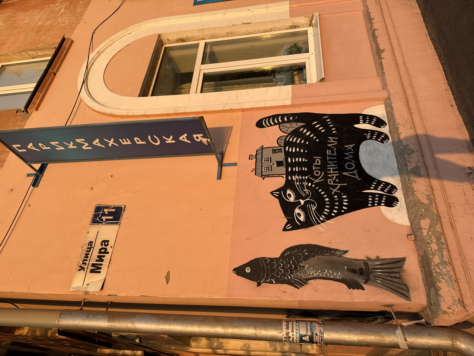 The image depicts a building in Votkinsk, Russia. The building is painted in a pinkish hue and has a blue sign with white Cyrillic letters, which translates to "Hairdresser". Beneath the sign is a street sign that reads "Ulitsa Mira 11", indicating the building is located at 11 Peace Street.
On the wall, there are cartoonish paintings of a black cat upside-down with a building on its belly and the phrase "Koty Hraniteli Doma" (Cats protectors of the home) written on it and a fish. Below the fish, there are some old stickers, one of which promotes a fair at 3-4 "Victory" Shopping Mall, which is held at Shkolnaya street.
The building also features windows with white frames. A metal pipe runs horizontally along the lower part of the building facade. The setting suggests a street scene with architectural elements and wall art providing local flavor.