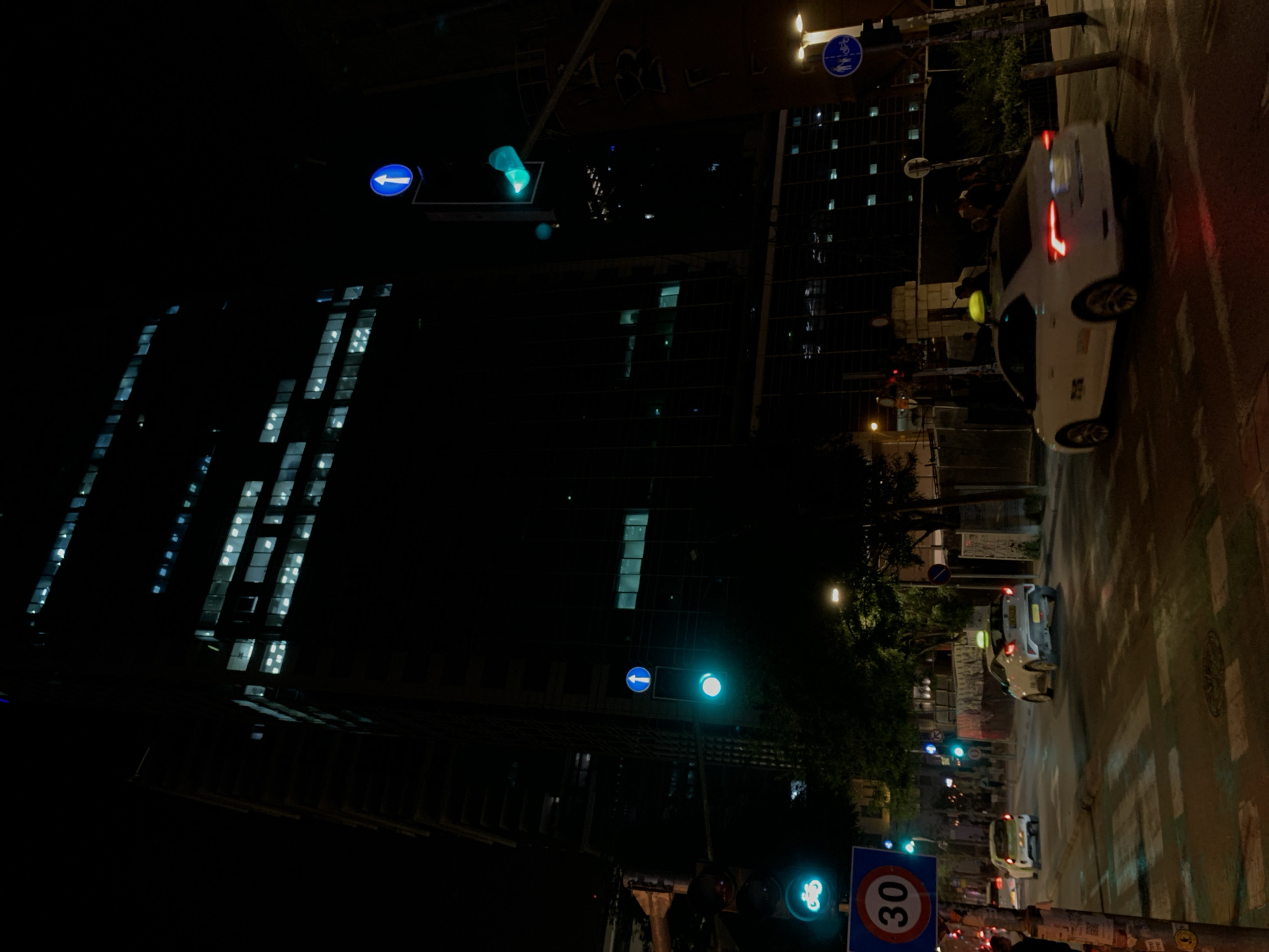 At night in Tel Aviv, Israel, a streetscape is dominated by tall buildings with illuminated windows on the left. A white car is turning on a wet street on the right. Traffic lights and street signs are visible, including a speed limit sign reading "30". The street is illuminated by streetlights and car headlights.