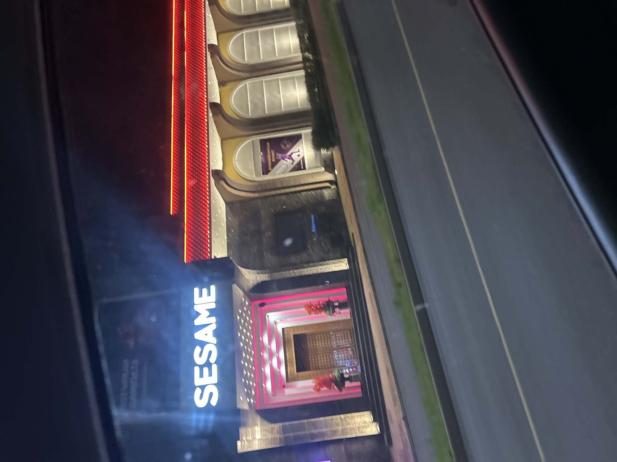 Captured from an elevated position, the image presents the "SESAME" building in Unknown, Unknown. The building is characterized by an illuminated vertical sign that spells out "SESAME," a decorative red lighting structure, and arched windows on the upper floors. The entryway is adorned with pink lighting and decorative plants. Alongside the building, a road runs, and the image appears to be taken from inside a vehicle, as indicated by the window frame visible on the sides. The time appears to be nighttime, judging by the building's illumination.
