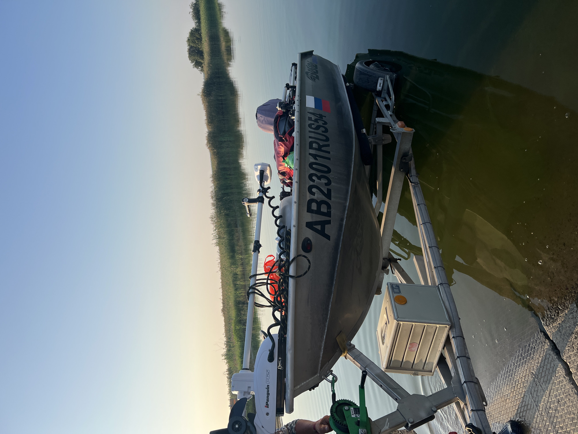 An aluminum boat, bearing the registration number AB2301RUS54 and a Russian flag decal, is positioned on a trailer partially submerged in the water of a calm lake. The boat is equipped with a white electric trolling motor labeled "iPenguin PG5". Red and black ropes are visible inside the boat, along with a covered object. The trailer has a green winch mechanism attached to its front. A metal box with a yellow reflector is mounted on the trailer's frame. The scene appears to be set in Zav'yalovo, Russia, evidenced by the water body and vegetation characteristic of the region. The sky is clear and blue, transitioning to a lighter hue near the horizon, suggesting either early morning or late afternoon. The water is green and reflects the boat, trailer, and surrounding reeds.