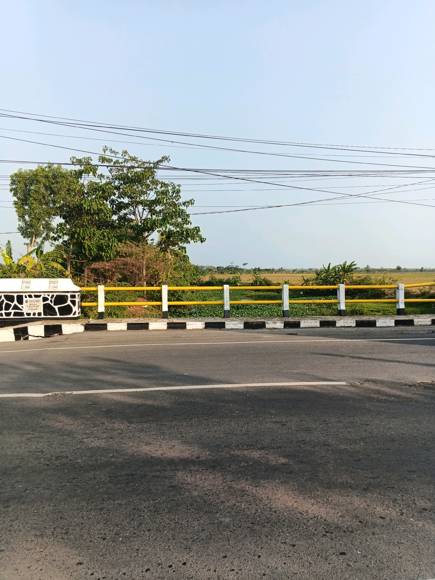 The image shows a roadside view in Doplang, Indonesia. The foreground is dominated by a dark asphalt road with a white line marking the lane. The middle ground features a white and black striped concrete barrier, followed by a yellow and white railing that runs along the edge of a body of water, possibly a canal or river, covered with green aquatic plants. Beyond the railing, there is a stretch of land with vegetation, including trees and what appears to be rice paddies or fields. Power lines are strung across the clear, light blue sky. A white concrete structure with a decorative black and white pattern is visible on the left side, with text inscribed on it. The scene appears to be captured during the day, with sunlight casting shadows on the road. There are no people or animals visible in the frame.