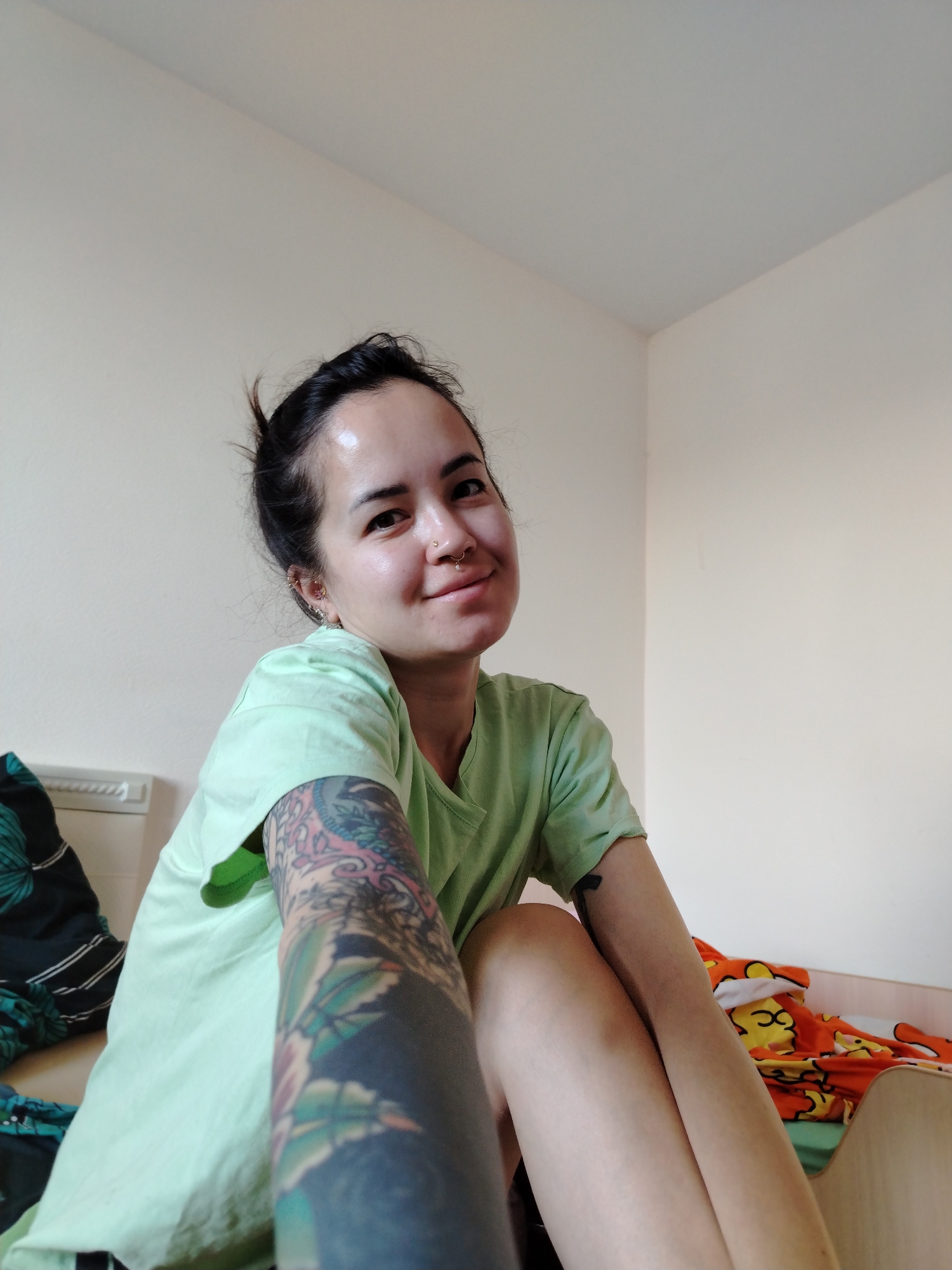 A woman with fair skin, dark hair pulled back into a bun, and multiple piercings in her nose and ears is sitting on a bed in an interior room. She is wearing a light green t-shirt and has a full sleeve tattoo on her left arm. The room has off-white walls. The bed has a patterned blanket. The location is Noviy, Russia.