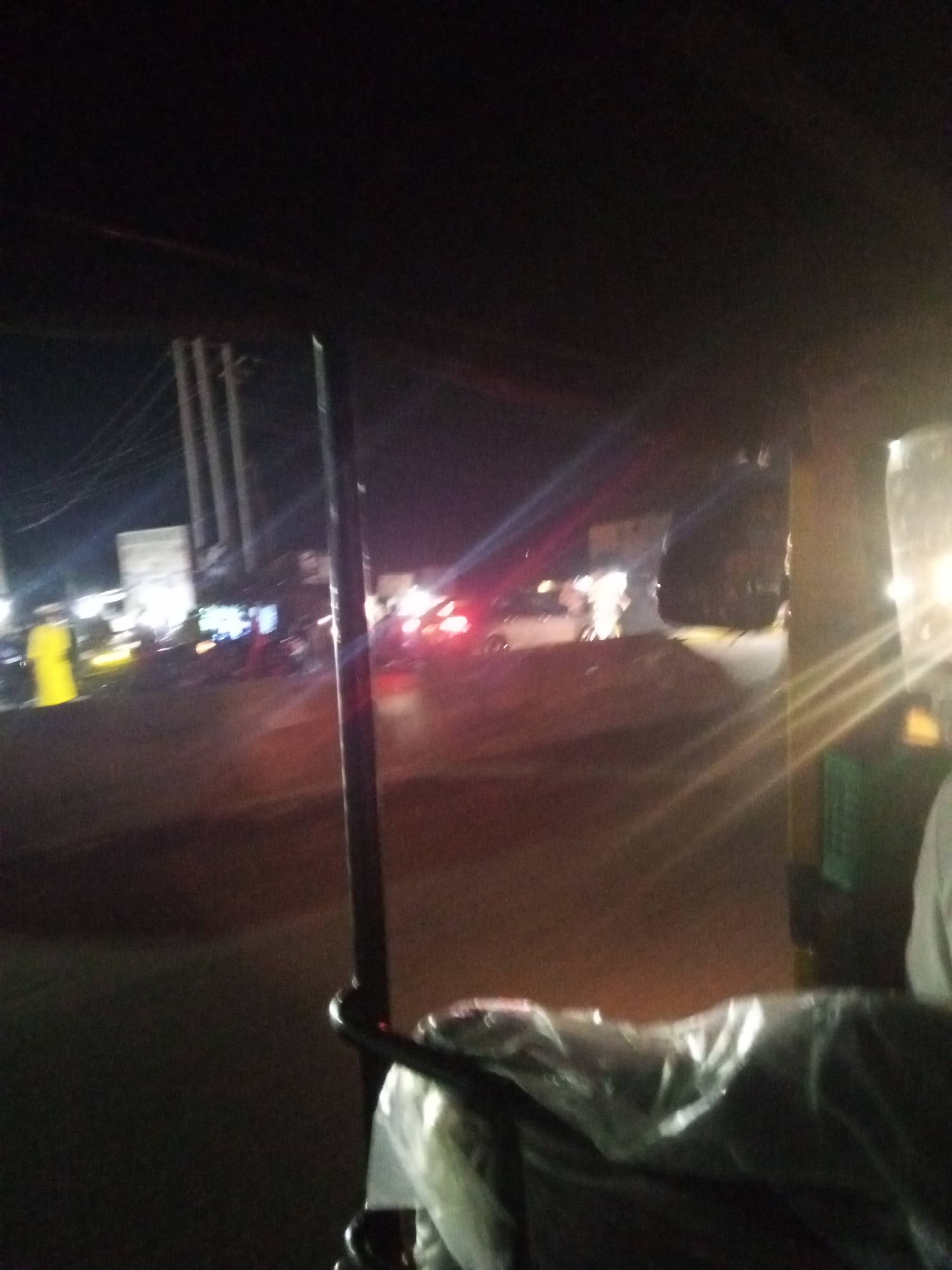The image depicts a nighttime scene on a street in Lokoja, Nigeria, viewed from inside an auto-rickshaw. The foreground is dominated by the interior of the auto-rickshaw, with a plastic-covered seat and a metal frame visible. The exterior scene is blurry and illuminated by various lights. Several vehicles, including a car with red taillights, are present on the road, suggesting traffic. Streetlights and lights from buildings can also be seen in the background. The overall impression is of a busy, urban environment at night.